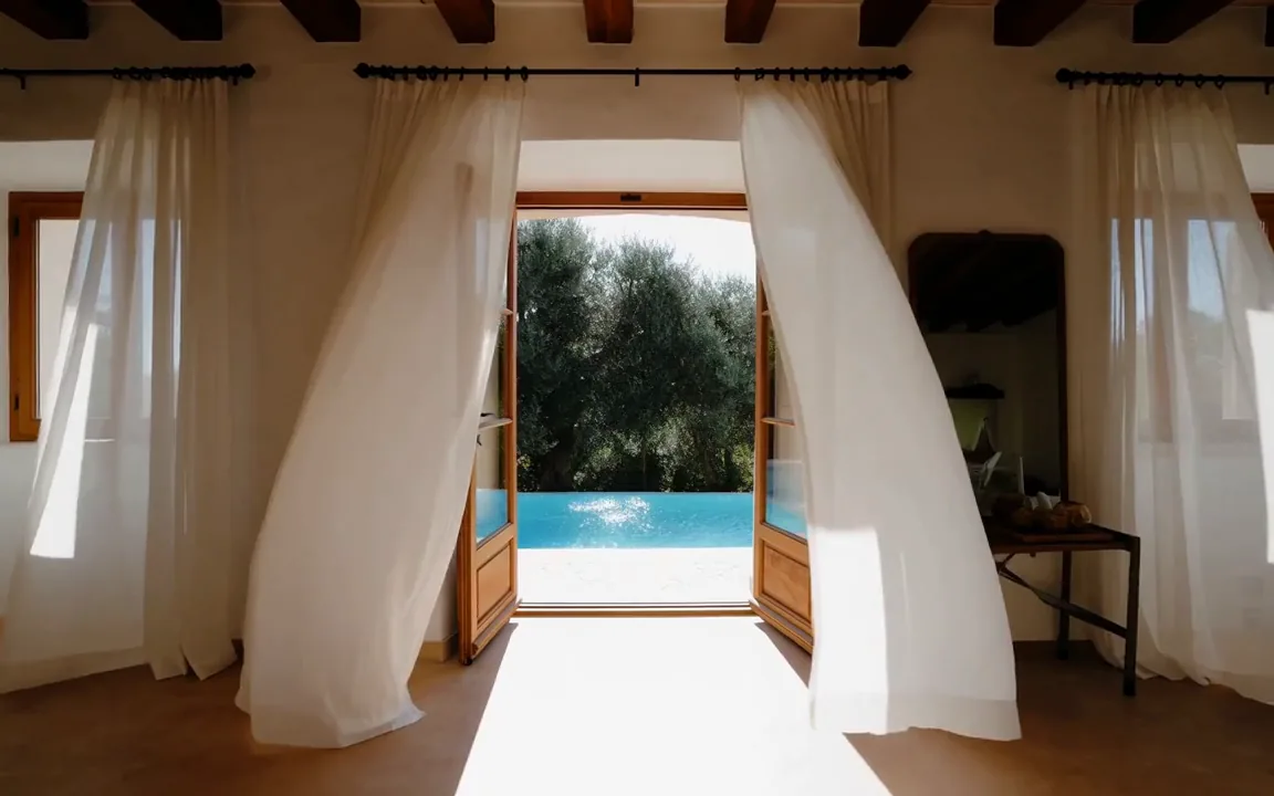 Camera pushing toward open terrace doors of a villa as the curtains lift and the pool glints