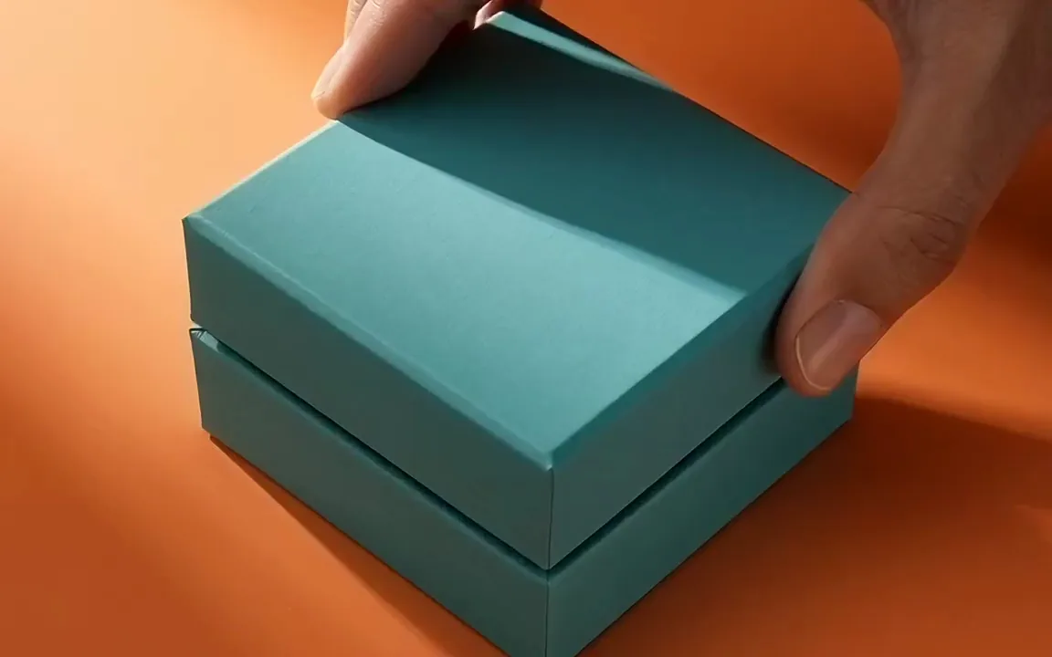 Text to video promo clip of a hand opening a teal gift box to reveal a gold necklace on pink silk