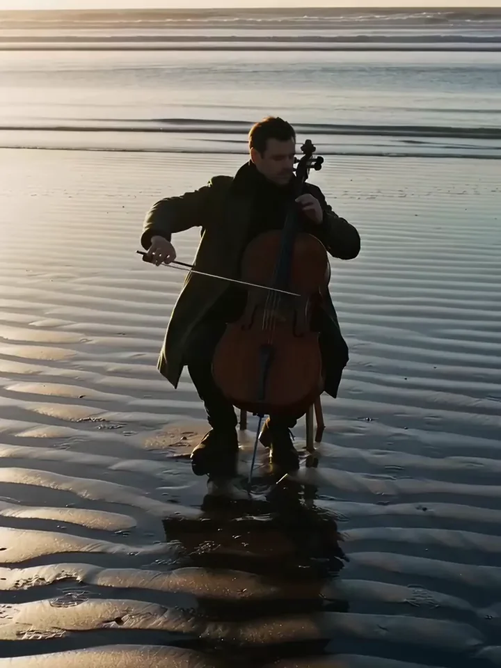 Un violoncellista che tiene una lunga nota con l'archetto, da solo su una spiaggia con la bassa marea