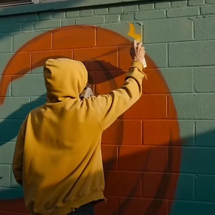 A street artist in a yellow hoodie spraying a yellow sunburst onto an orange mural on a teal wall