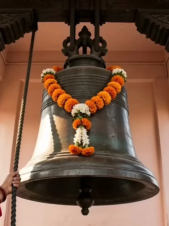 A bronze temple bell hung with a marigold garland swinging beneath a carved wooden beam