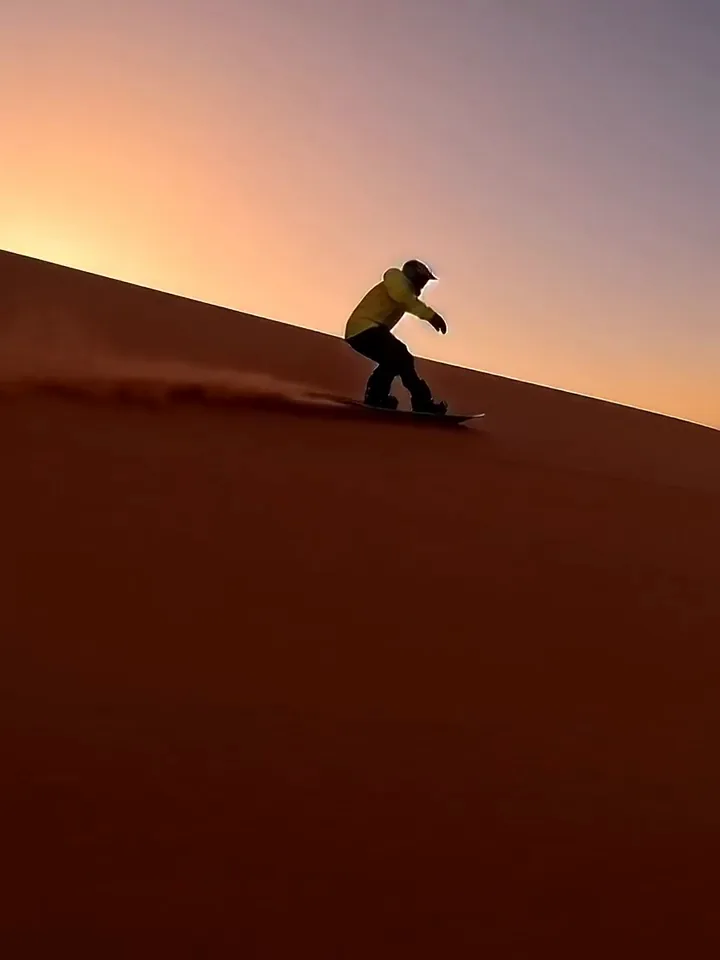 Sandboarder riding down a red dune at sunset, throwing a fan of sand