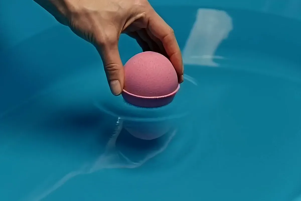 FLUX 3 video of a hand setting a pink bath bomb on blue water as it fizzes and blooms magenta clouds