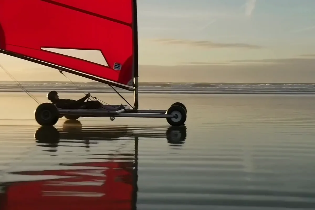A sand yacht with a red sail tipping up on one side in a gust on a wet beach