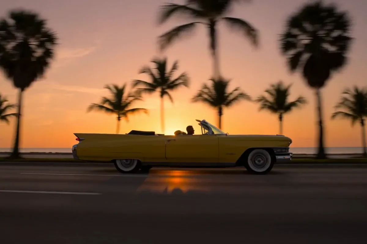 Text to video clip of a yellow vintage convertible cruising past palm trees at sunset