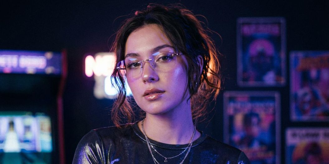 Portrait of a woman wearing glasses in neon-lit room with arcade and posters in the background.