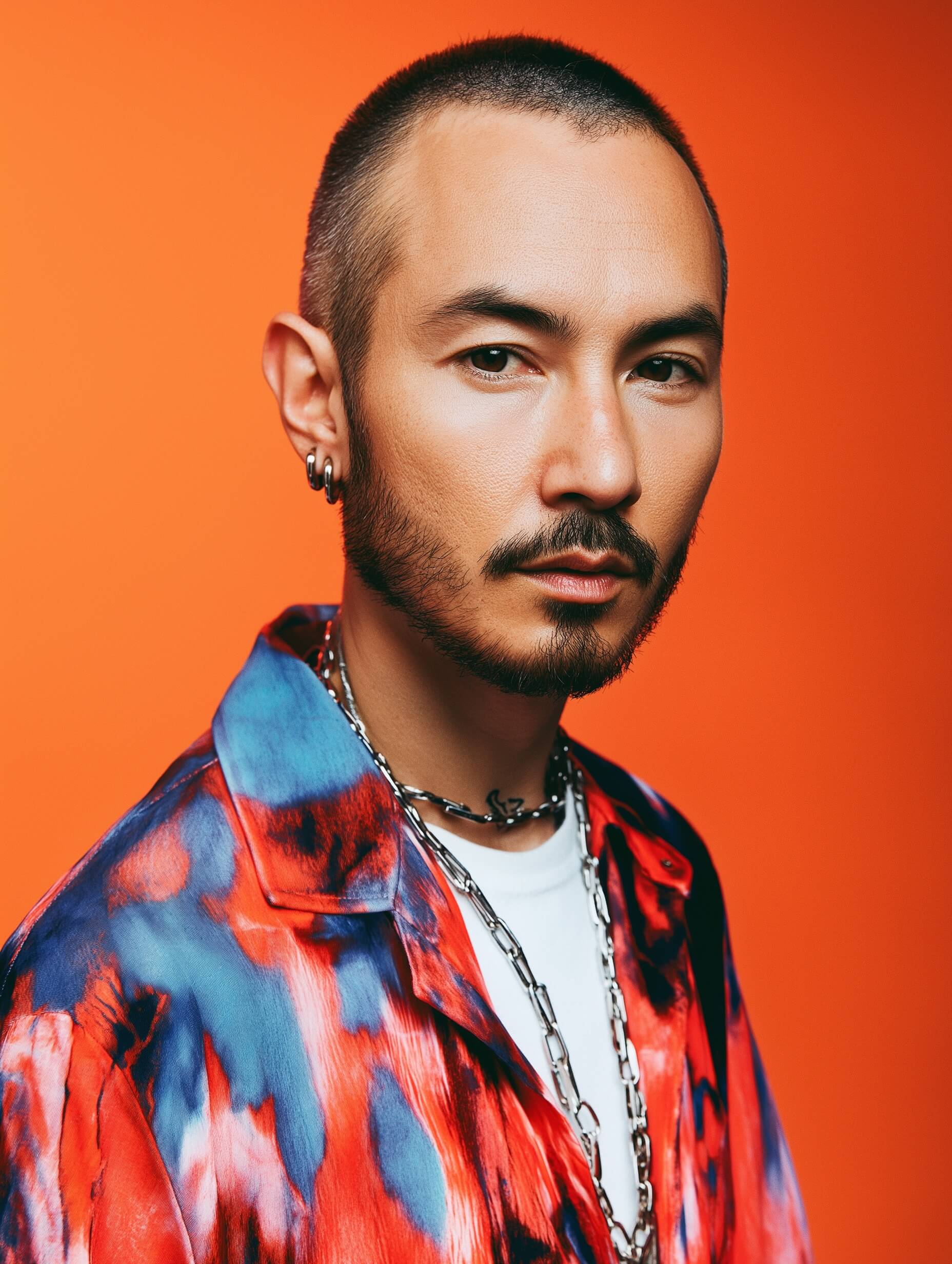 An intriguing portrait captures a man against a solid orange background, gazing intently at the camera. He sports a close buzz cut, a neat mustache, and a goatee, dressed in a stylish shirt featuring bold, abstract patterns in red, blue, and black, unbuttoned over a white shirt. A chain necklace accentuates his outfit, adding subtle elegance. Photographed with soft, even lighting to flatter his features, the portrait's composition is tightly framed to focus on his face, while a portrait lens creates a sharp image with a beautifully blurred backdrop, highlighting the vibrant color contrast and modern urban style.