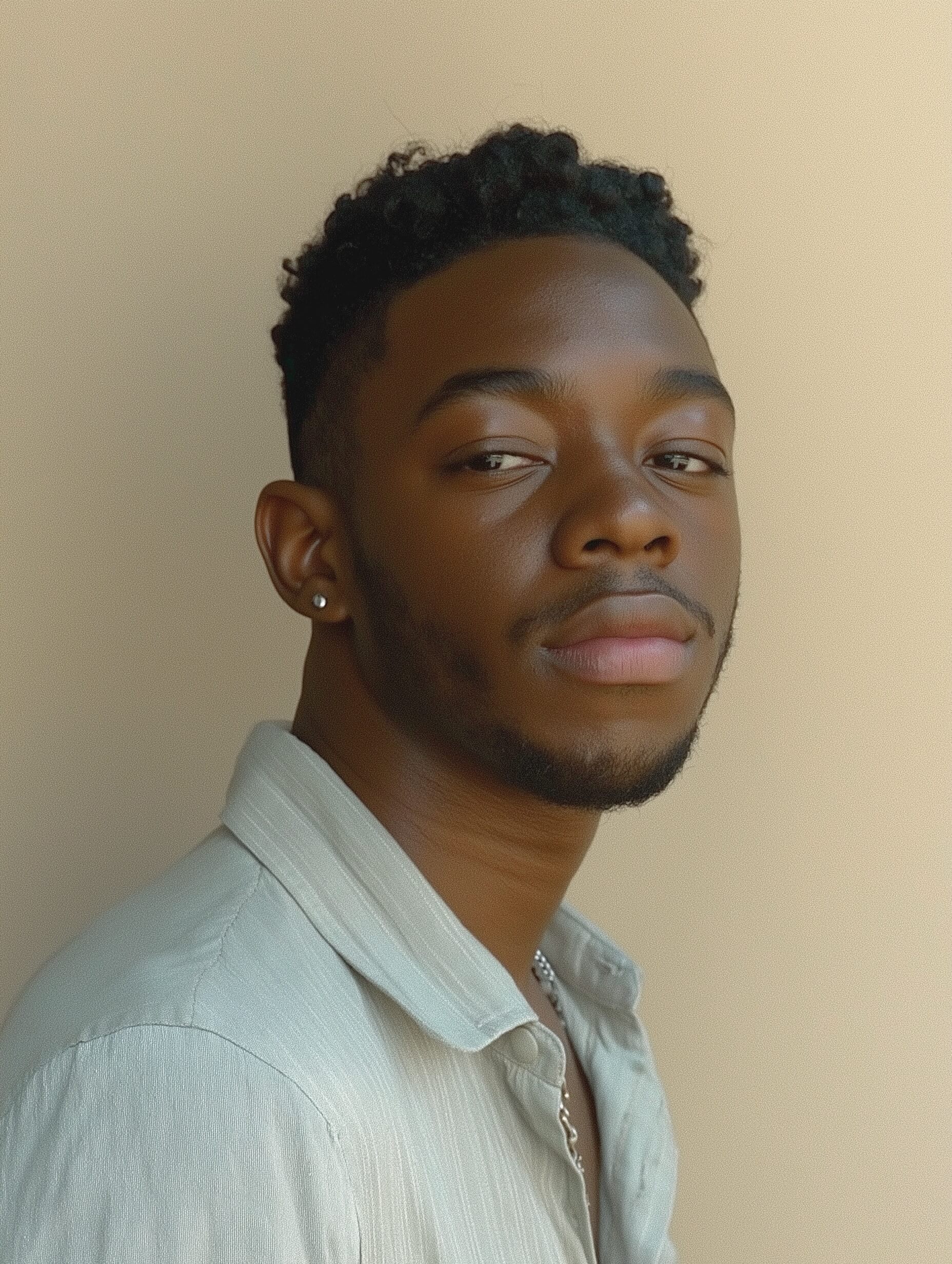 In this serene portrait, a young Black man with a focused expression wears a light gray, short-sleeve button-up shirt over his relaxed frame. With minimal styling, the soft gray tones of his shirt gently contrast the warm beige background. The lighting is soft and evenly distributed, avoiding harsh shadows by using a large diffused light source positioned in front. The image likely employs a mid-range aperture (f/4 - f/5.6) for sharp focus and soft background, captured by a 50mm or 85mm lens. Framed with the subject’s upper torso centered, the earthy color palette creates a warm feel, enhanced slightly in post-processing. The overall minimalist composition emphasizes simplicity and highlights the man’s natural features.