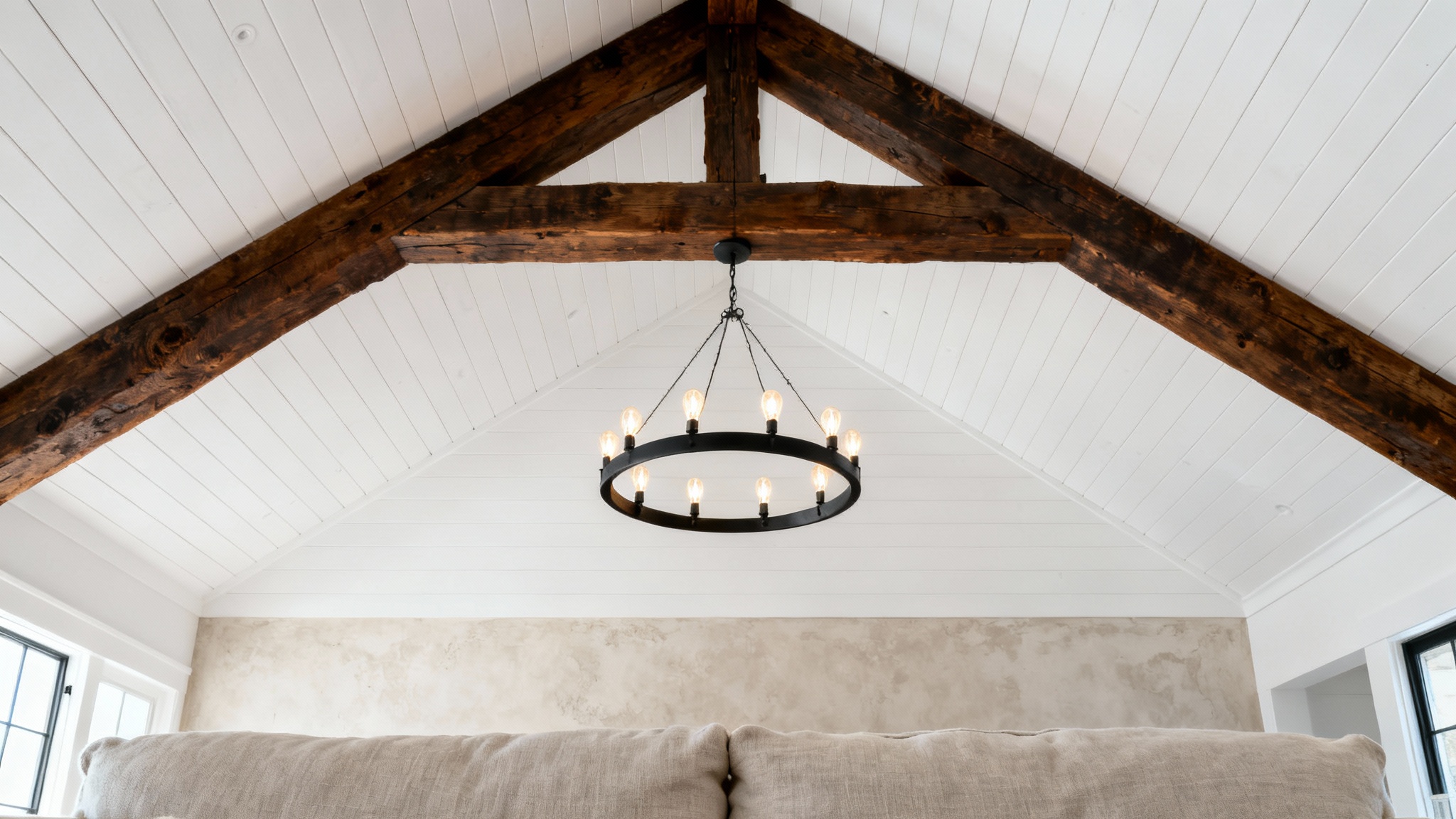 A low-angle shot of a modern farmhouse vaulted ceiling with dark wood beams, white shiplap, and a large black iron chandelier, conveying a sense of spaciousness and rustic elegance.