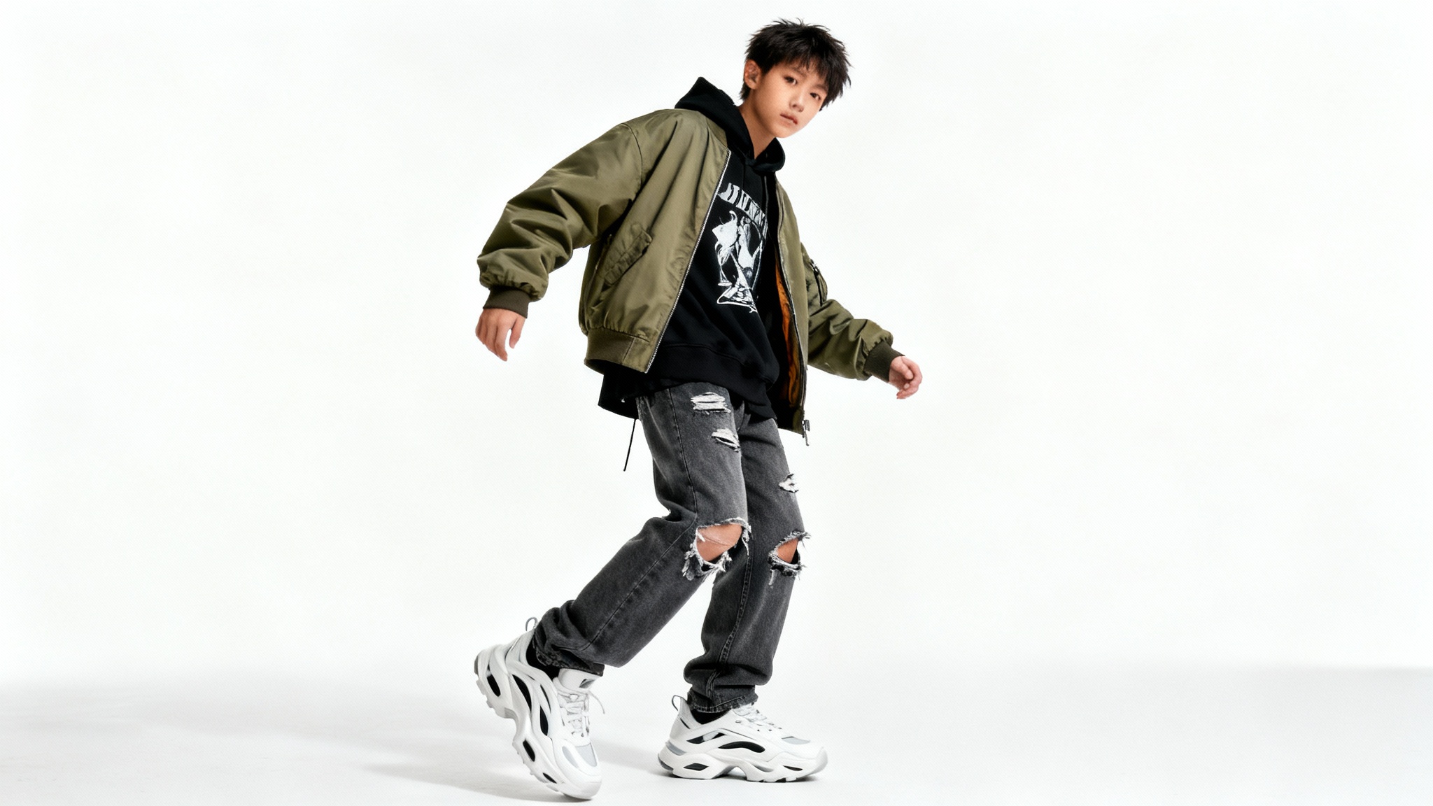A full-body studio photograph of a person modeling a contemporary urban fashion outfit, including a hoodie, bomber jacket, and jeans, isolated against a solid white background.