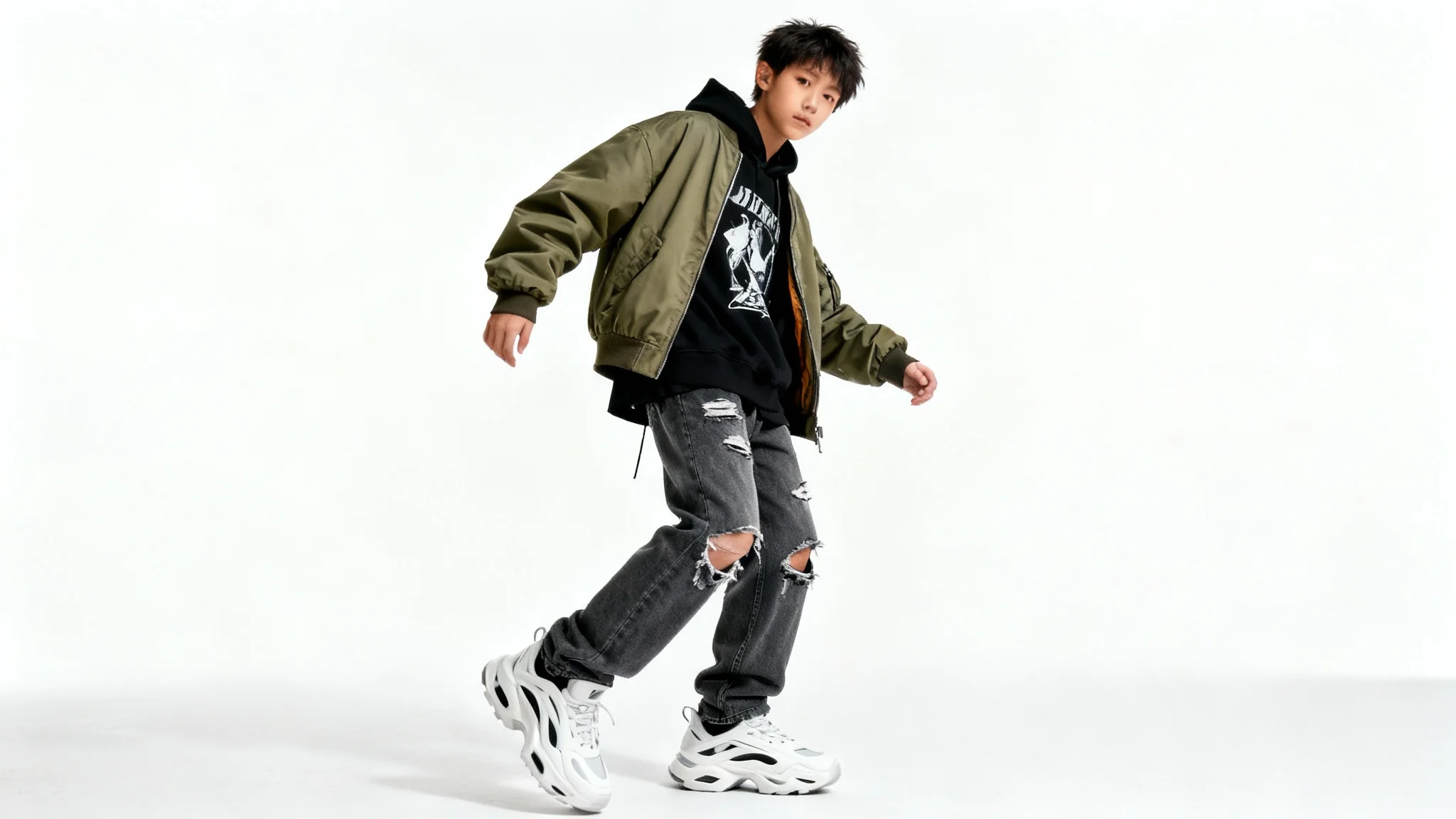 A full-body studio photograph of a person modeling a contemporary urban fashion outfit, including a hoodie, bomber jacket, and jeans, isolated against a solid white background.