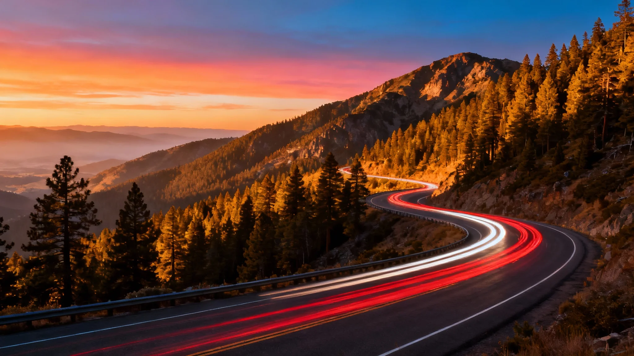 An aerial photograph of a winding highway through mountains at sunset, with long exposure light trails from cars creating streaks of red and white light.