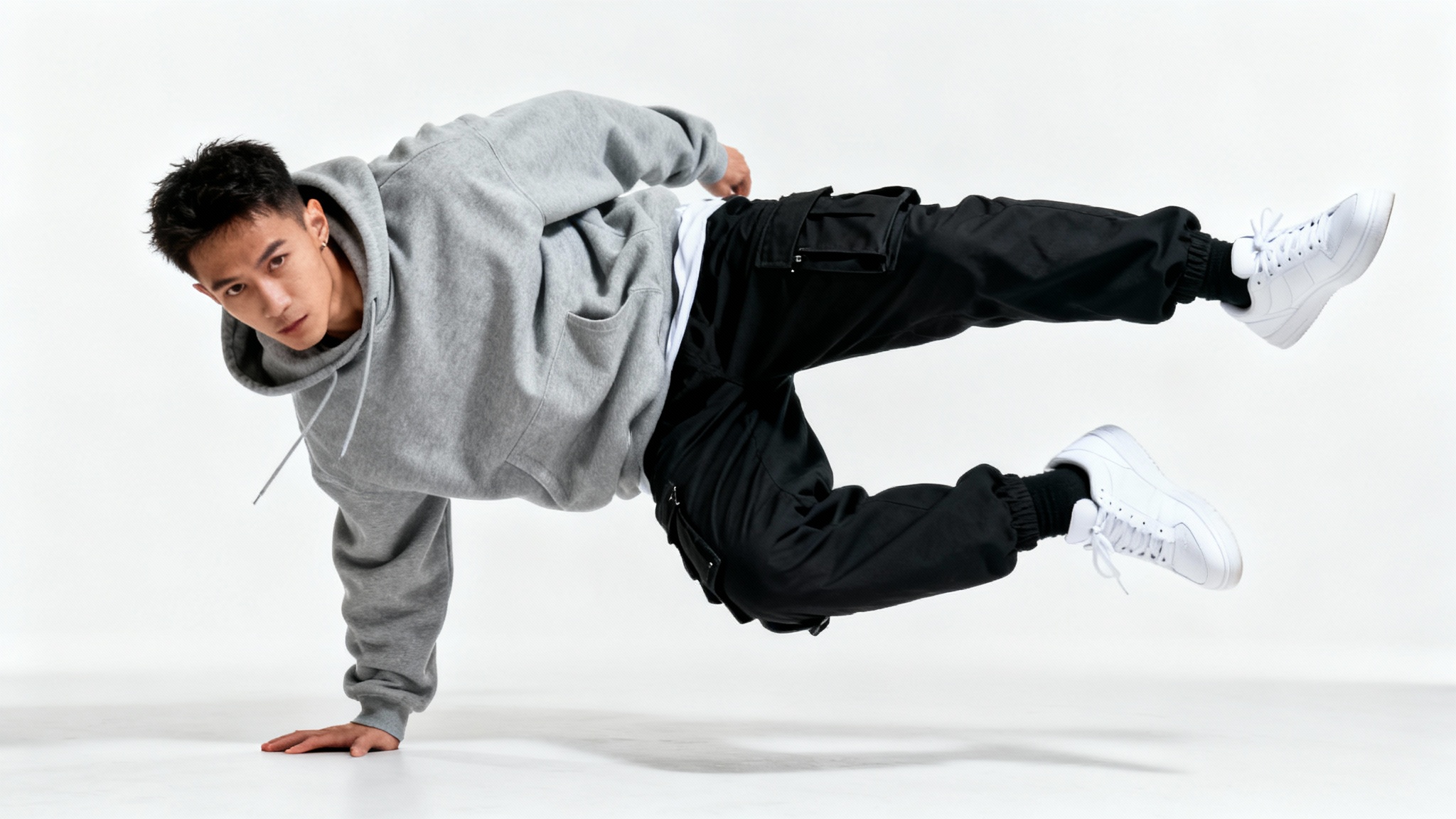 A male street dancer in a hoodie and cargo pants is frozen mid-pose, balancing on one hand against a plain white background.