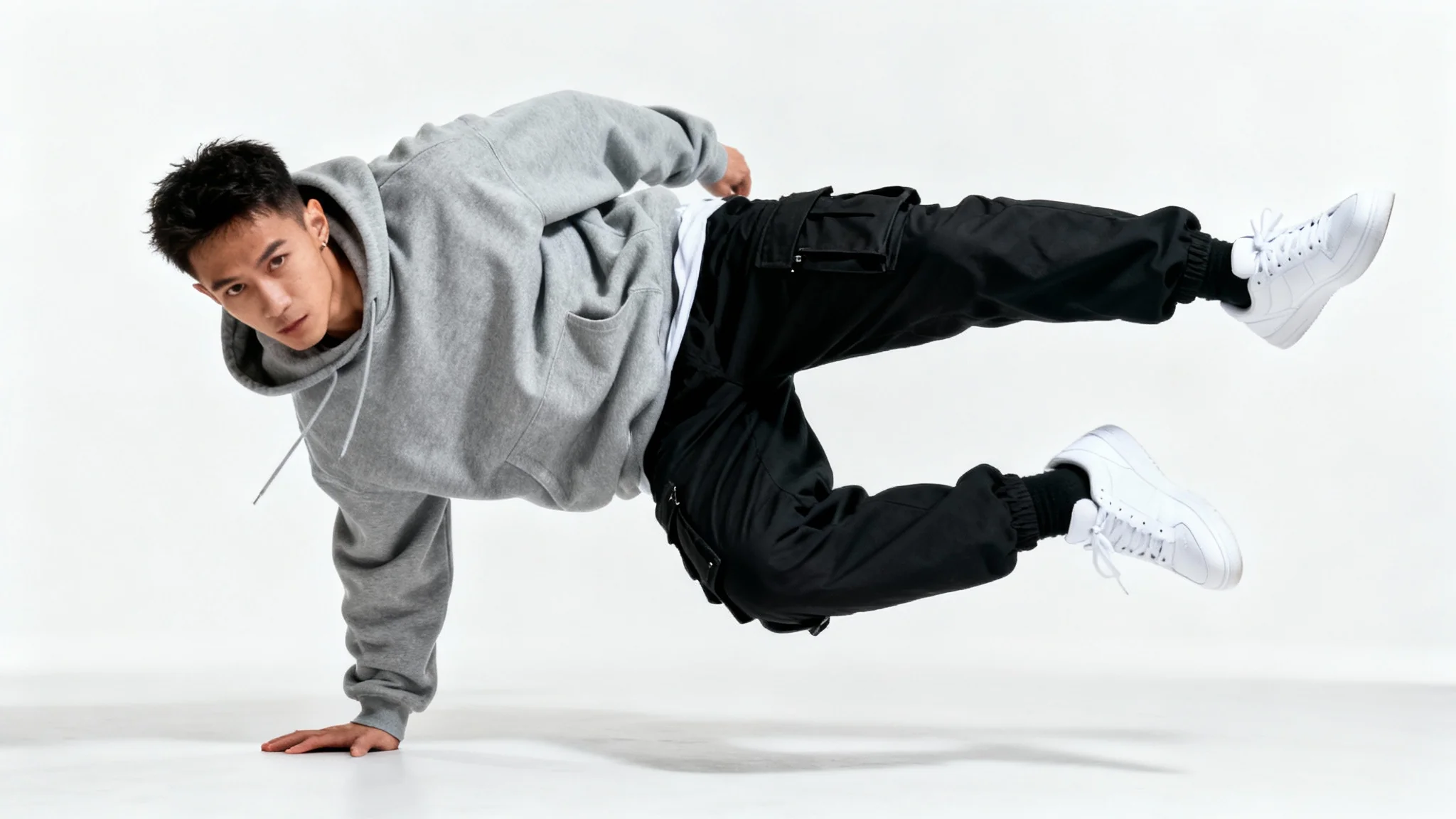 A male street dancer in a hoodie and cargo pants is frozen mid-pose, balancing on one hand against a plain white background.