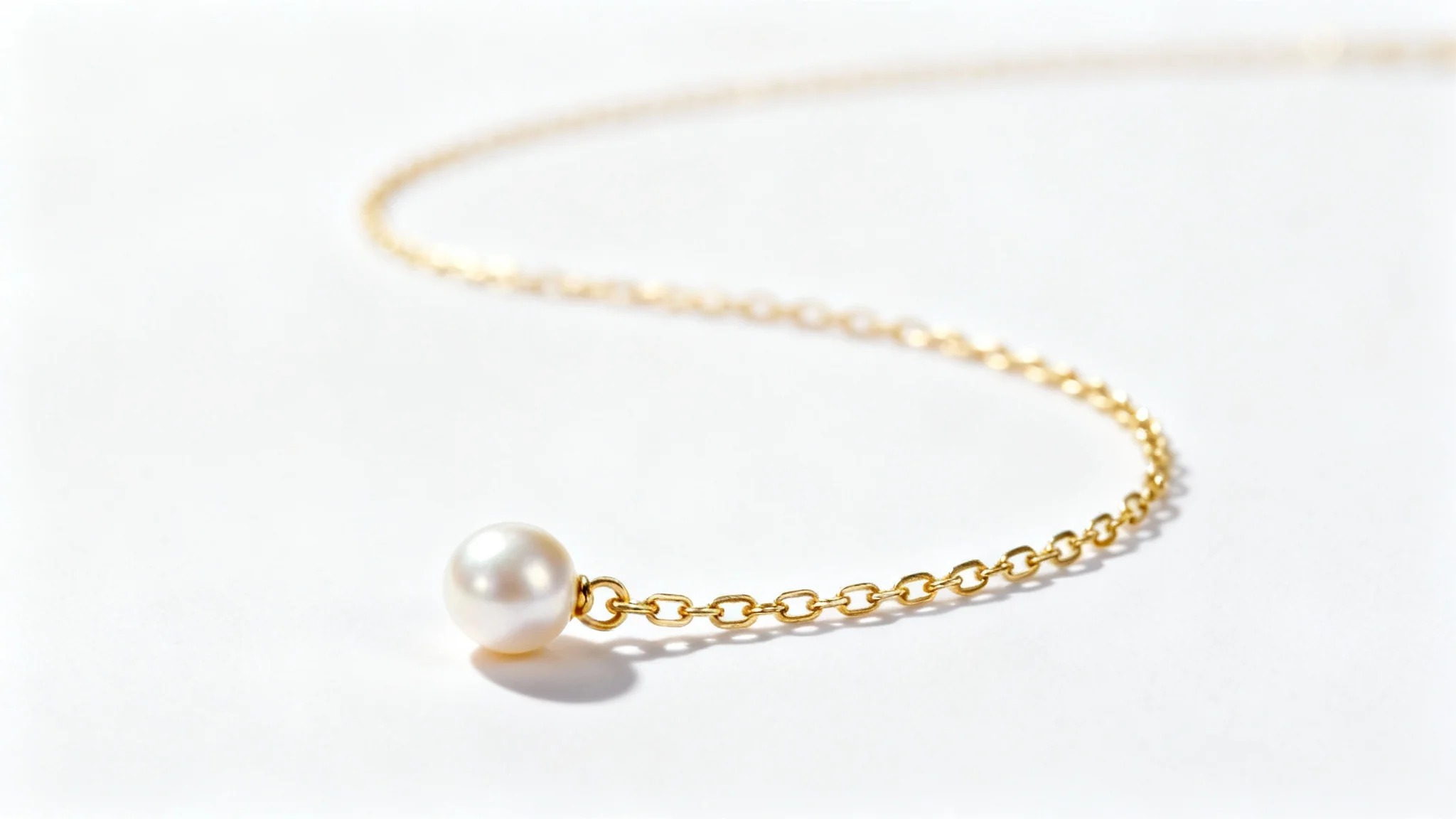 A high-end, professional product photograph of a delicate gold necklace with a single pearl, shot in a minimalist style on a stark white background.