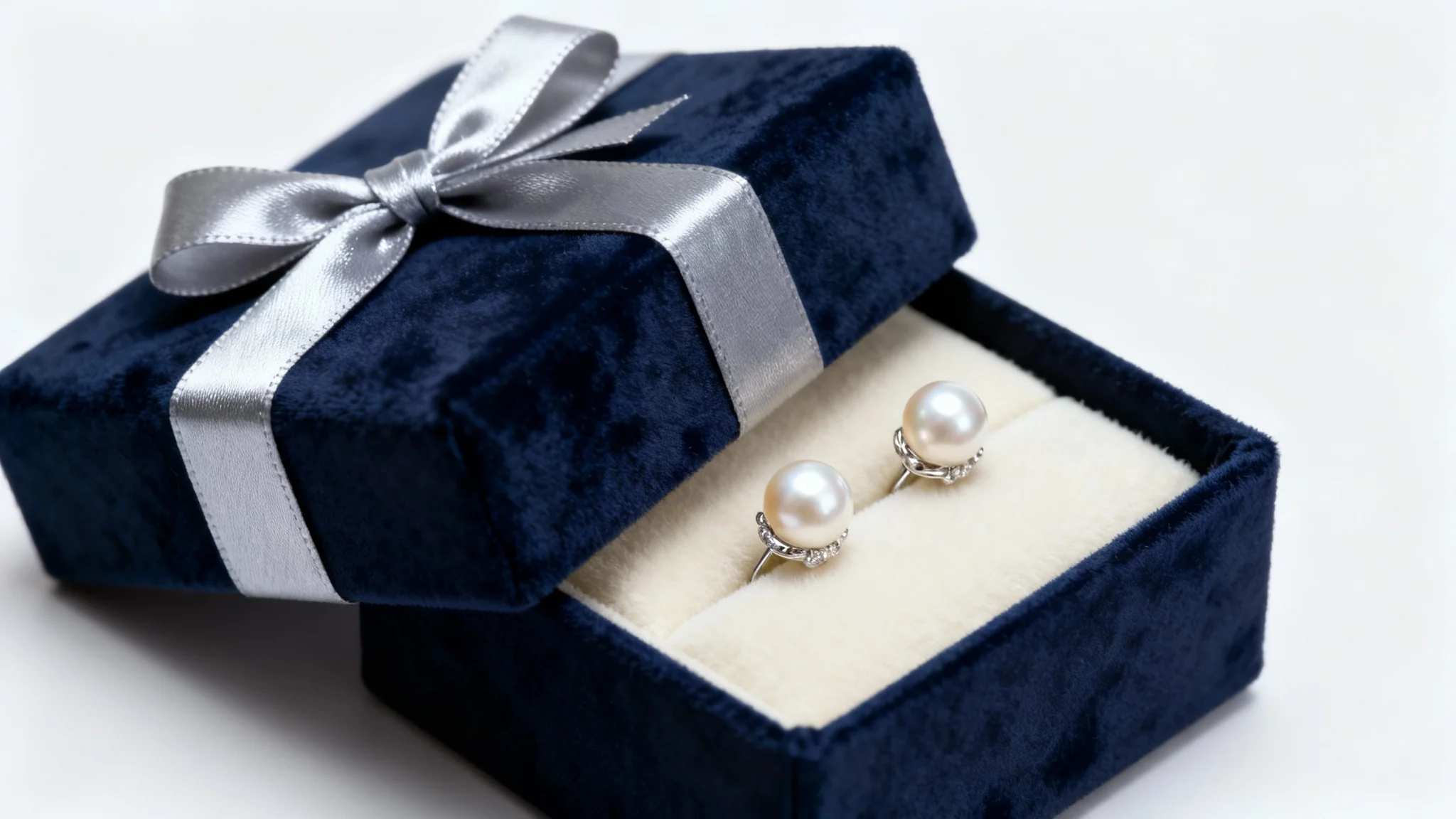 A luxurious navy blue jewelry gift box with a silver ribbon, slightly open to show a pair of pearl earrings on a cream velvet interior, set against a clean white background.