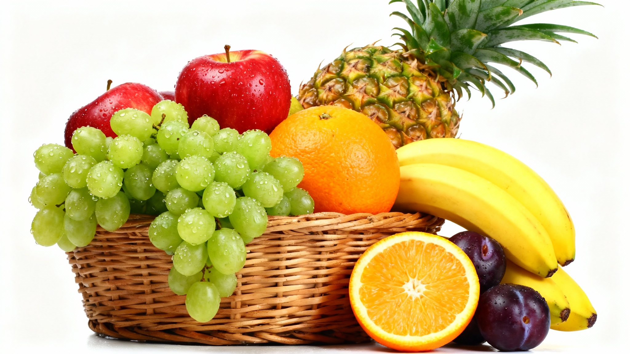 A studio photograph of a rustic wicker basket filled to the brim with a variety of fresh fruits, including apples, grapes, a pineapple, and bananas, set against a plain white background.