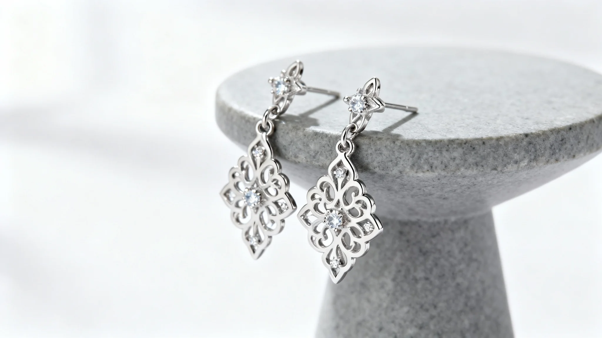 A professional studio photograph of a pair of elegant silver filigree earrings with diamonds, displayed on a grey stone pedestal against a clean white background.