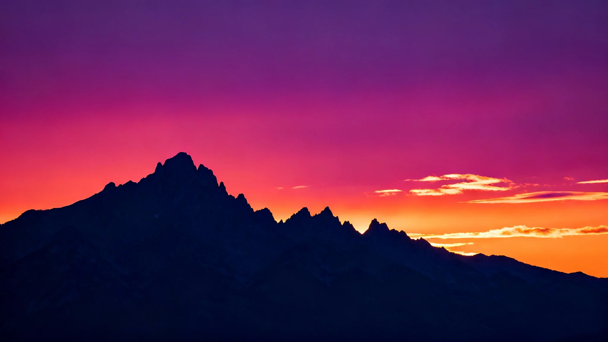 A serene and majestic silhouette of a jagged mountain range set against a vibrant sunset sky transitioning from orange to purple.