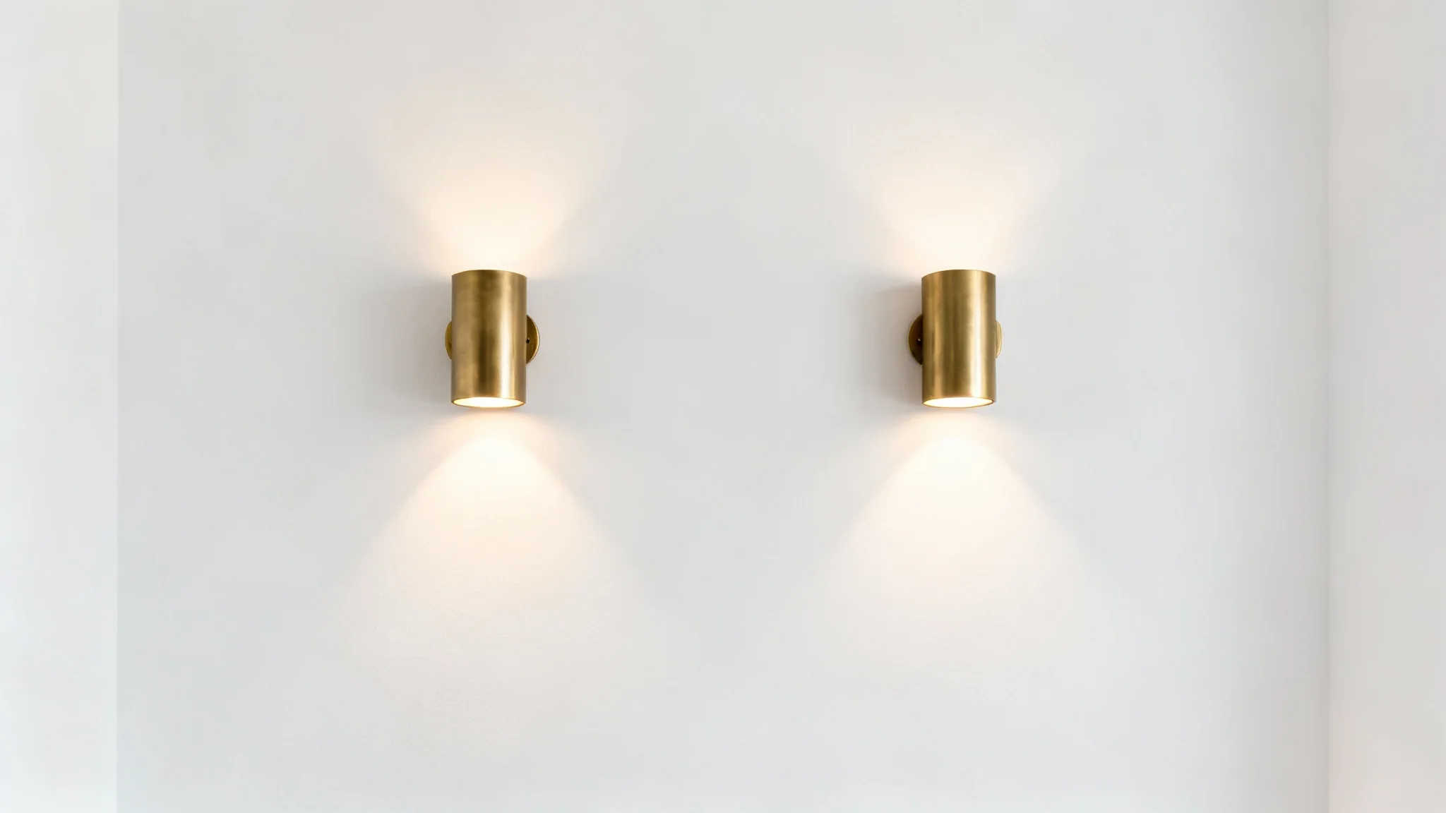 A pair of modern brushed brass wall sconces symmetrically mounted on a clean white wall, illuminated and casting a warm, ambient glow.