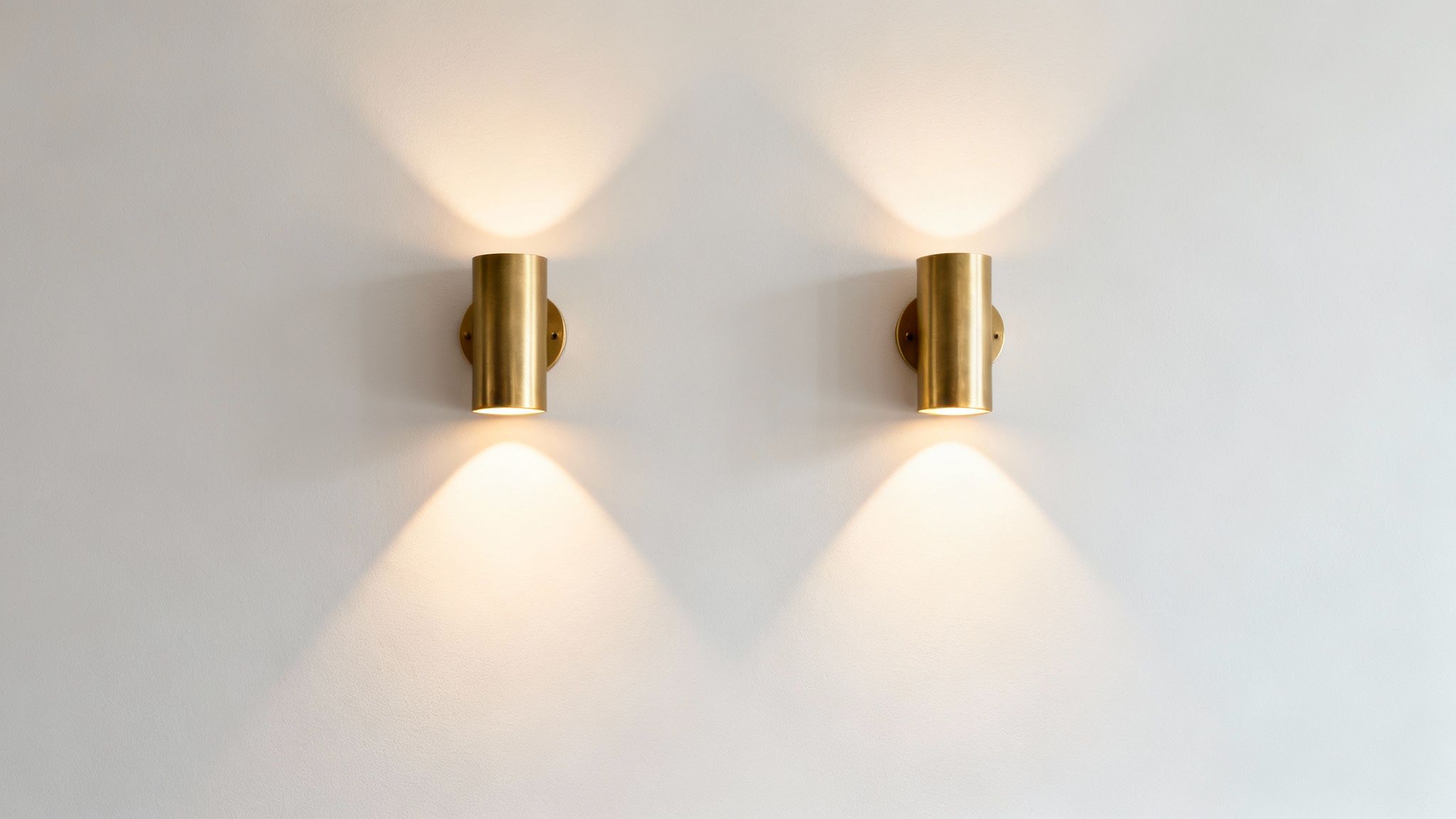 A photorealistic image of two modern, brushed brass wall sconces mounted on a clean off-white wall, casting a warm, elegant light.