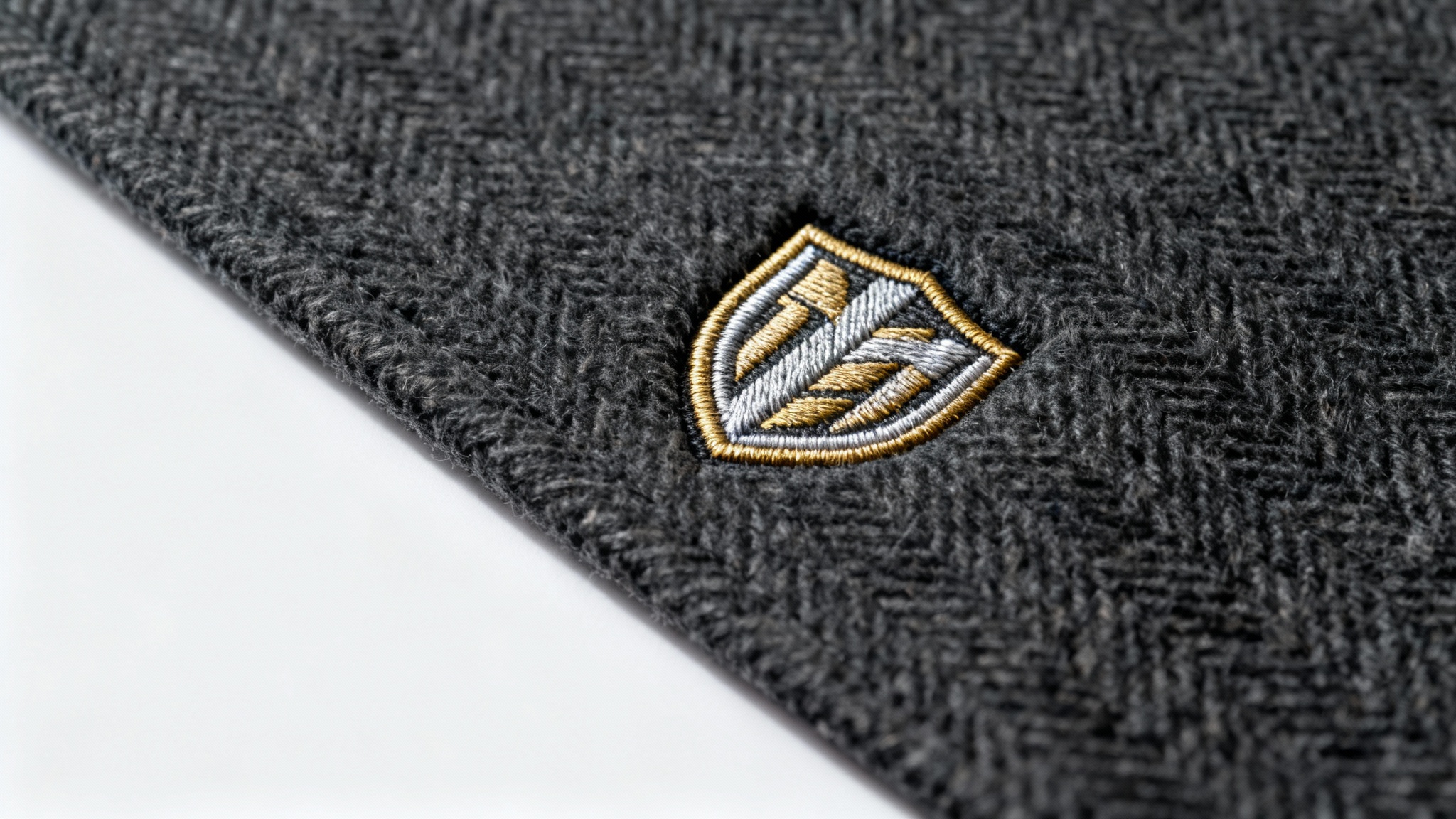 A close-up, photorealistic mockup of a metallic gold and silver embroidered crest logo on a piece of dark grey herringbone fabric, set against a clean white background.