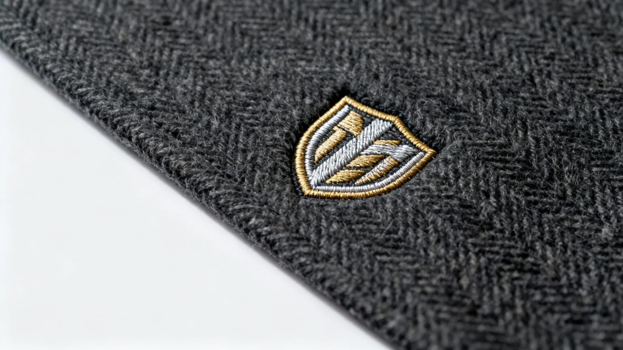 A close-up, photorealistic mockup of a metallic gold and silver embroidered crest logo on a piece of dark grey herringbone fabric, set against a clean white background.