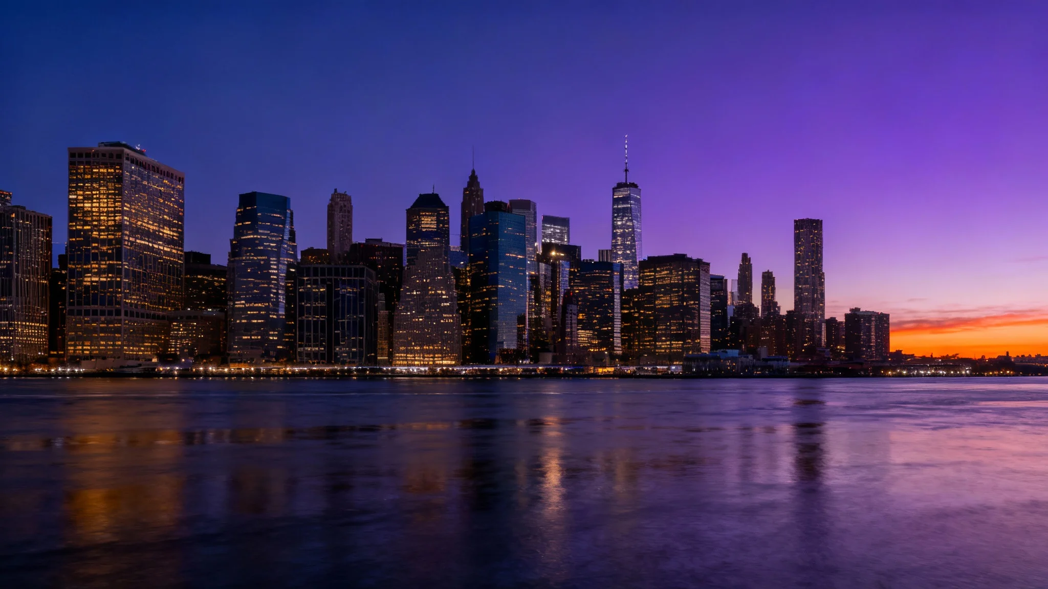 A panoramic photo of a modern city skyline at dusk, where glowing skyscrapers and a purple and orange sky are reflected in a wide river.