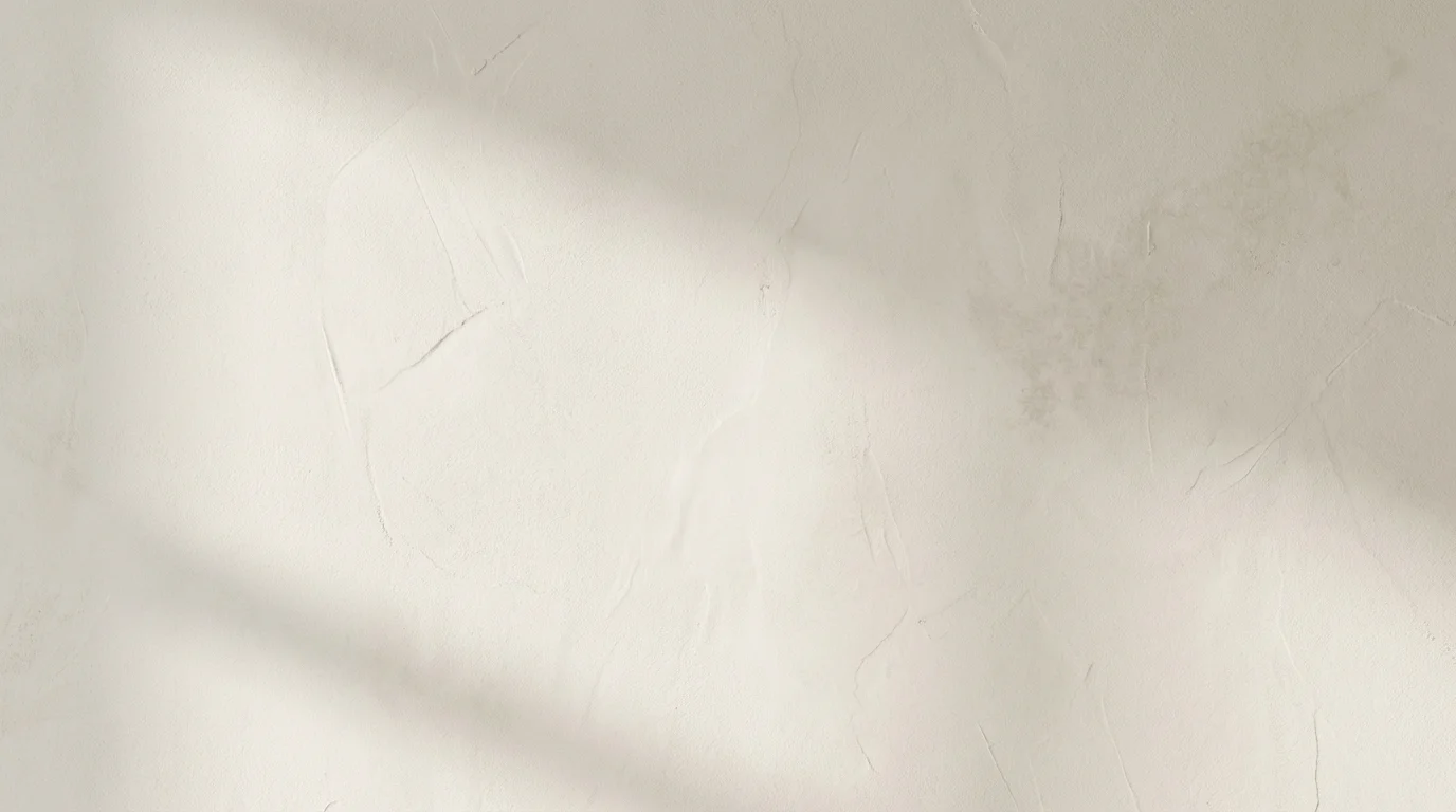 A detailed, macro shot of a warm off-white microcement wall texture. The surface is matte and smooth, with subtle variations highlighted by soft side lighting, creating a modern and minimalist look.