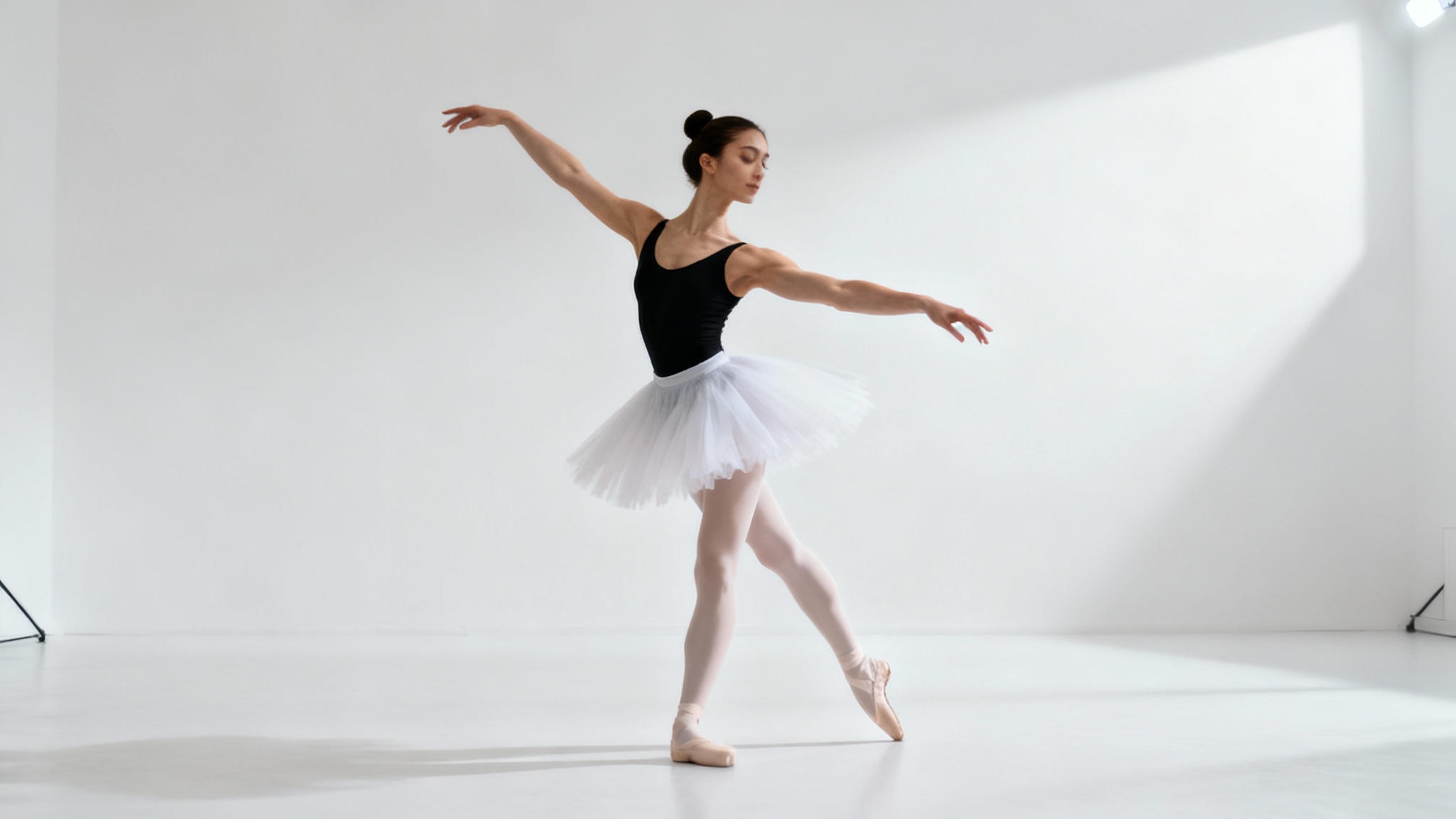 A full-body image of a female ballet dancer in a black leotard and white tutu, captured mid-pose against a stark white background.