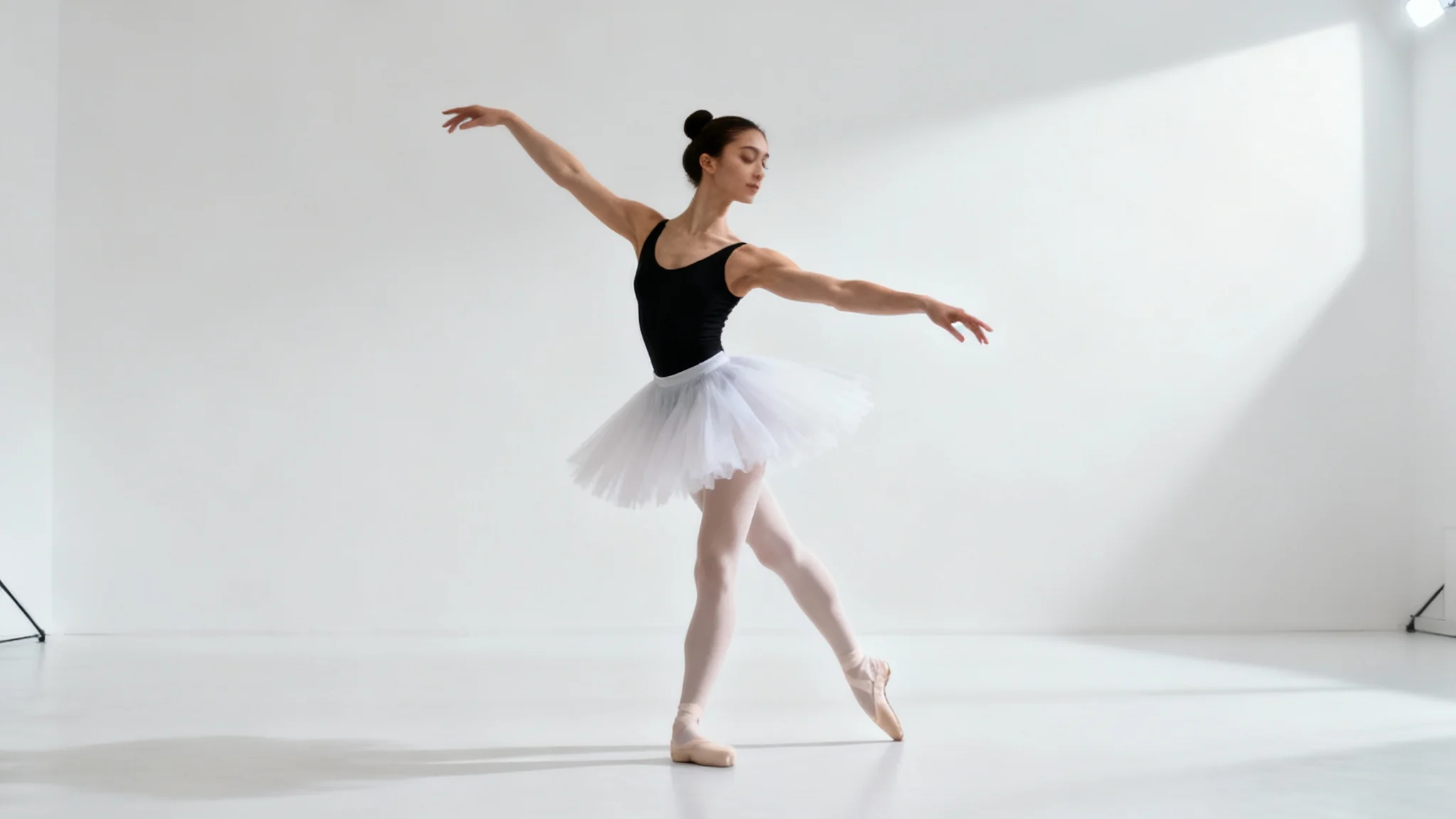 A full-body image of a female ballet dancer in a black leotard and white tutu, captured mid-pose against a stark white background.