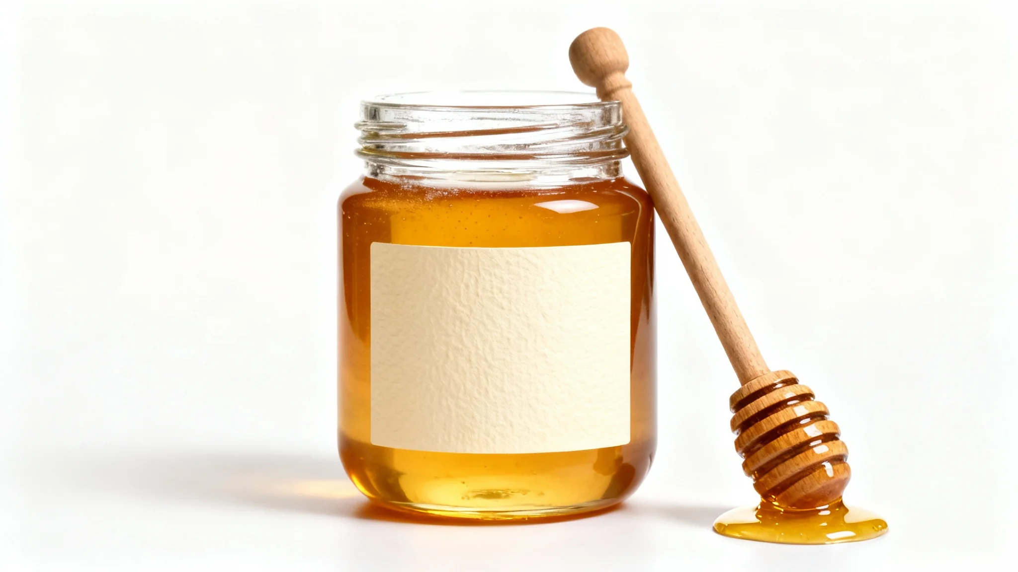 A photorealistic mockup of a glass honey jar filled with golden honey, featuring a blank paper label for branding. A wooden honey dipper with a drop of honey rests against it on a clean white background.