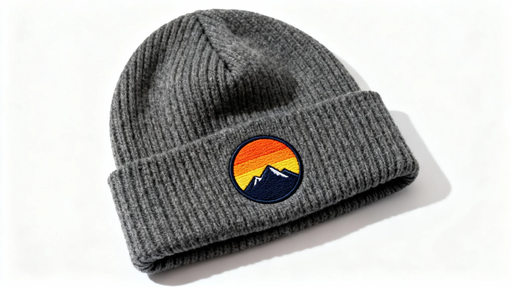 A close-up product shot of a charcoal grey knit beanie with a custom embroidered patch of a mountain sunset on its cuff, displayed against a clean white background.