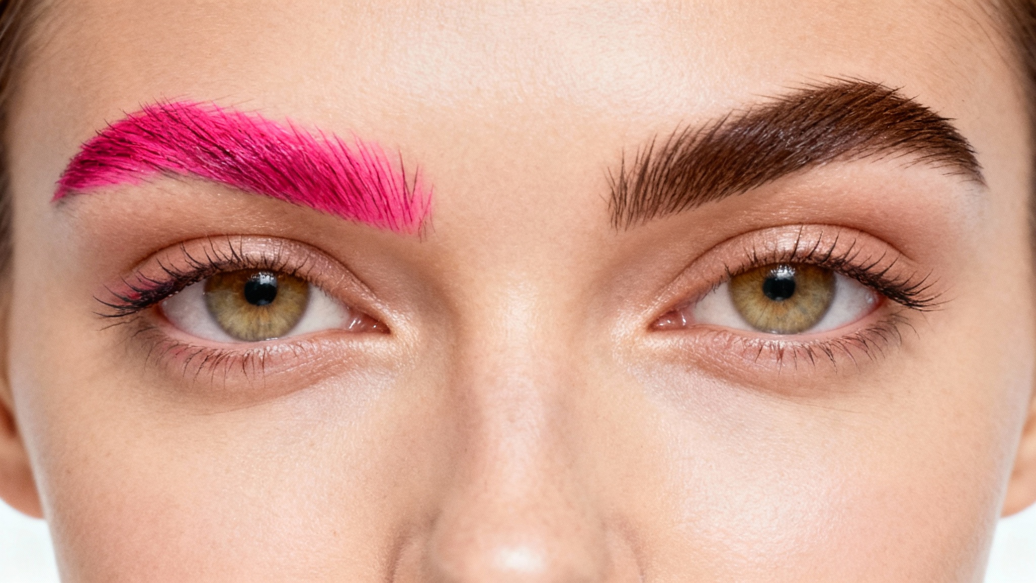 An extreme close-up beauty shot of a woman's eyes. One of her eyebrows is a natural dark brown, and the other is a vibrant pink, demonstrating an eyebrow color change effect, all against a stark white background.