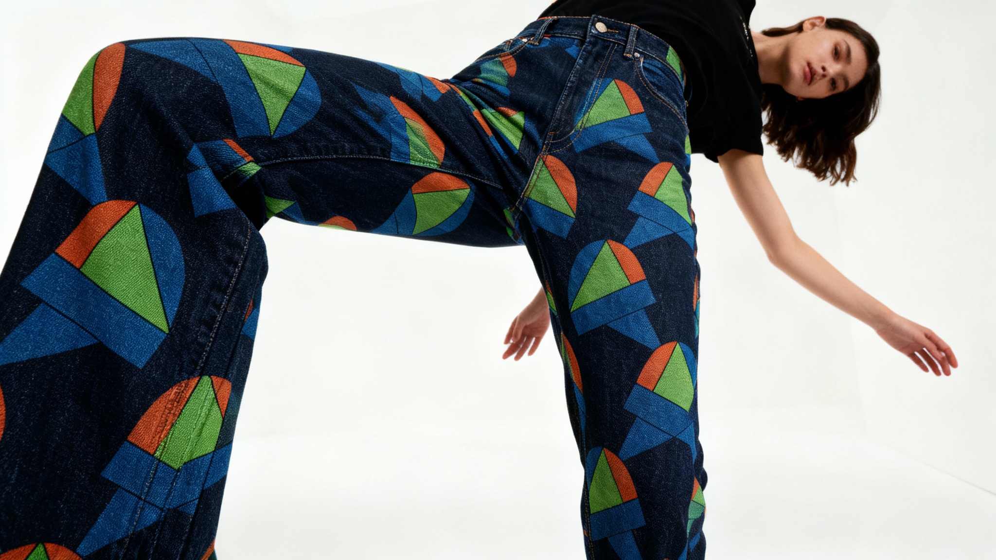 A close-up of a person wearing custom dark denim jeans featuring a vibrant, eclectic pattern of blue, green, and orange geometric and abstract shapes, shown against a minimalist white background.