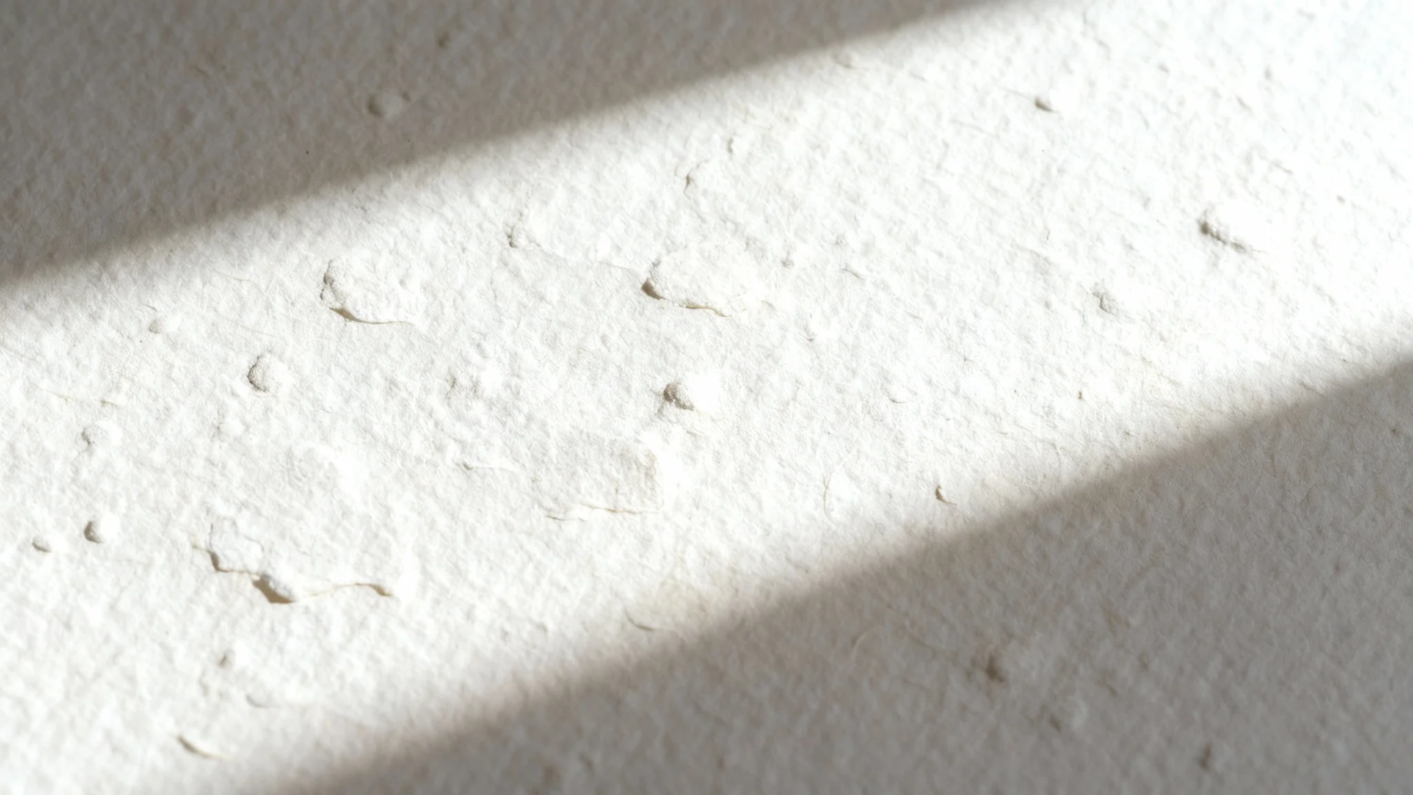 A detailed, macro close-up of a textured, off-white watercolor paper, with side lighting emphasizing its fibrous, bumpy surface against a white background.