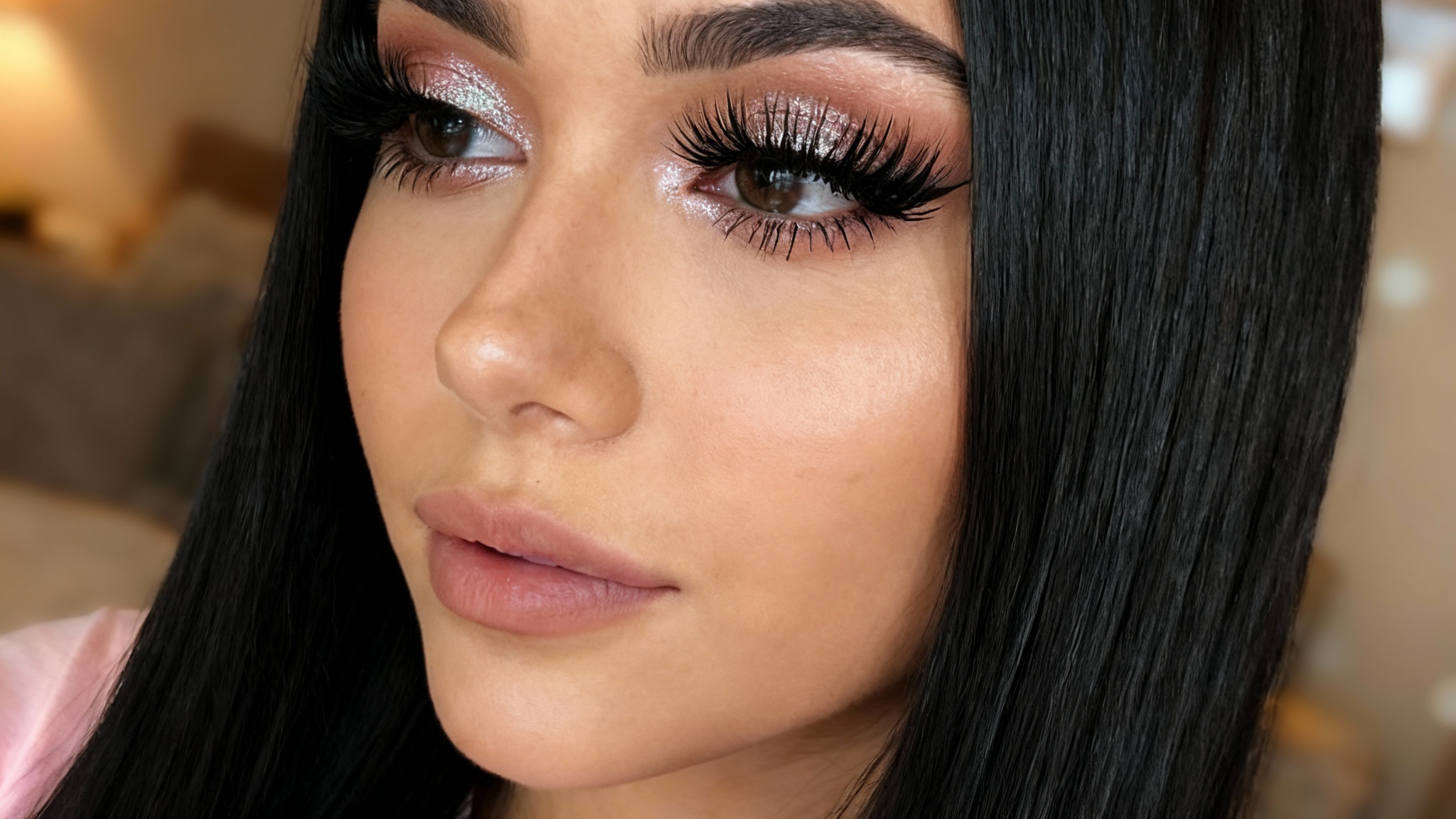 A close-up beauty shot of a woman's face, focusing on her eyes which have been enhanced with very long, full, beautiful eyelashes. She has dark hair and is looking slightly away from the camera with a soft expression.