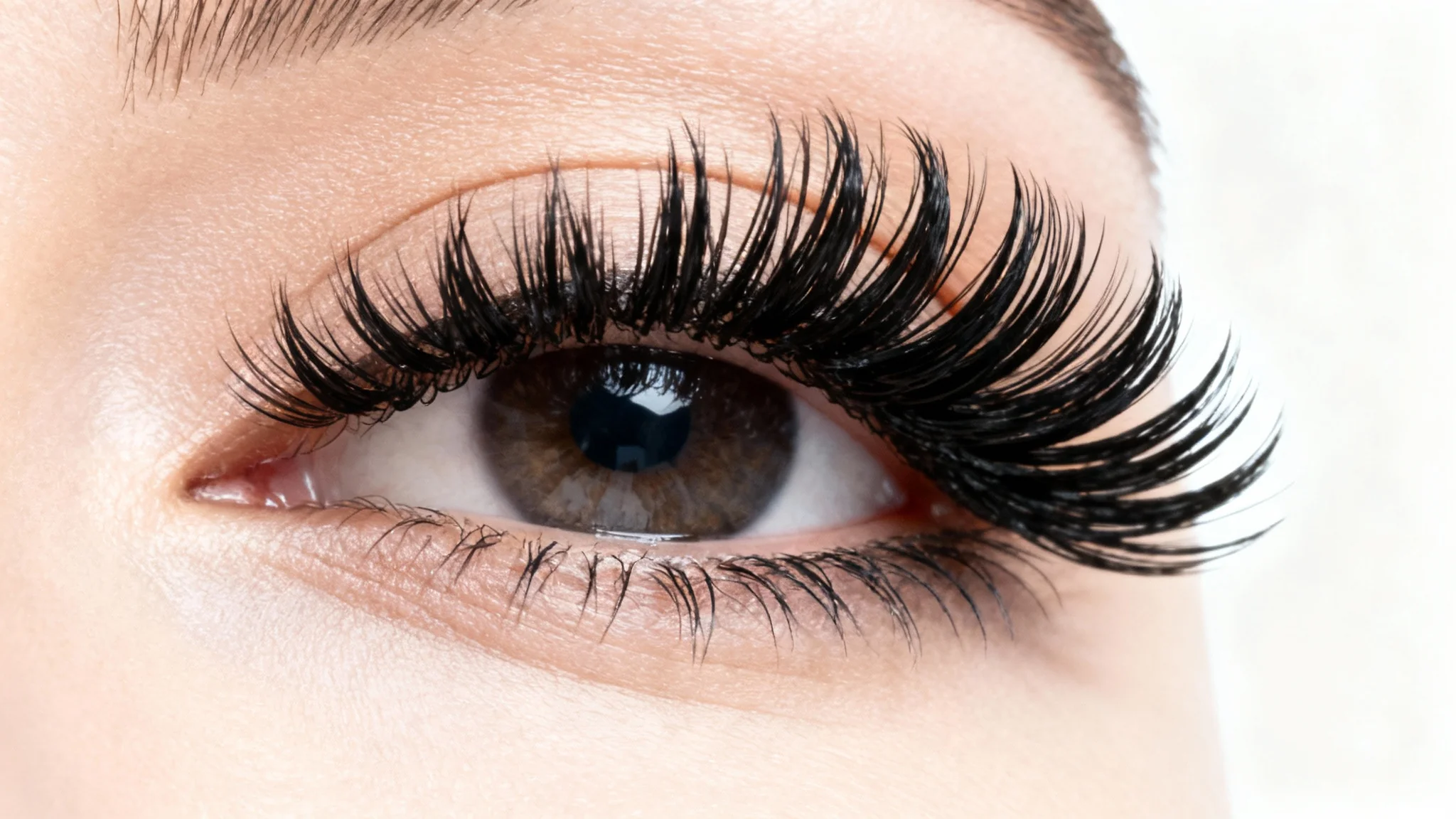 A close-up macro photo of a woman's eye, beautifully framed by long, voluminous black false eyelashes, set against a clean white background.