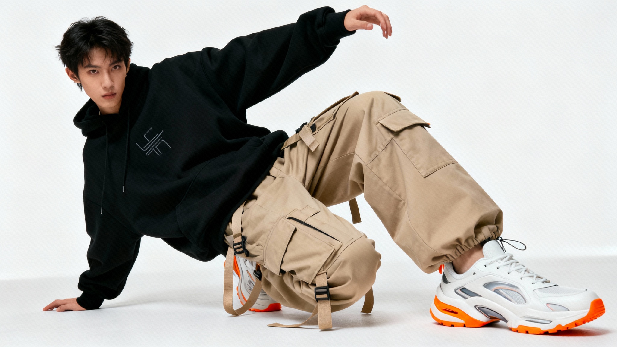 A full-body studio photograph of a person modeling a contemporary streetwear outfit, consisting of a black hoodie, beige cargo pants, and white sneakers, against a plain white background.