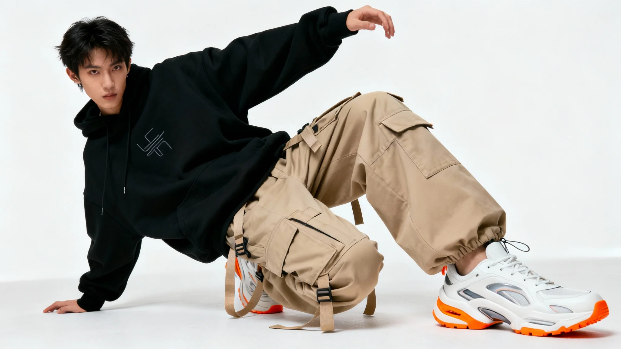 A full-body studio photograph of a person modeling a contemporary streetwear outfit, consisting of a black hoodie, beige cargo pants, and white sneakers, against a plain white background.