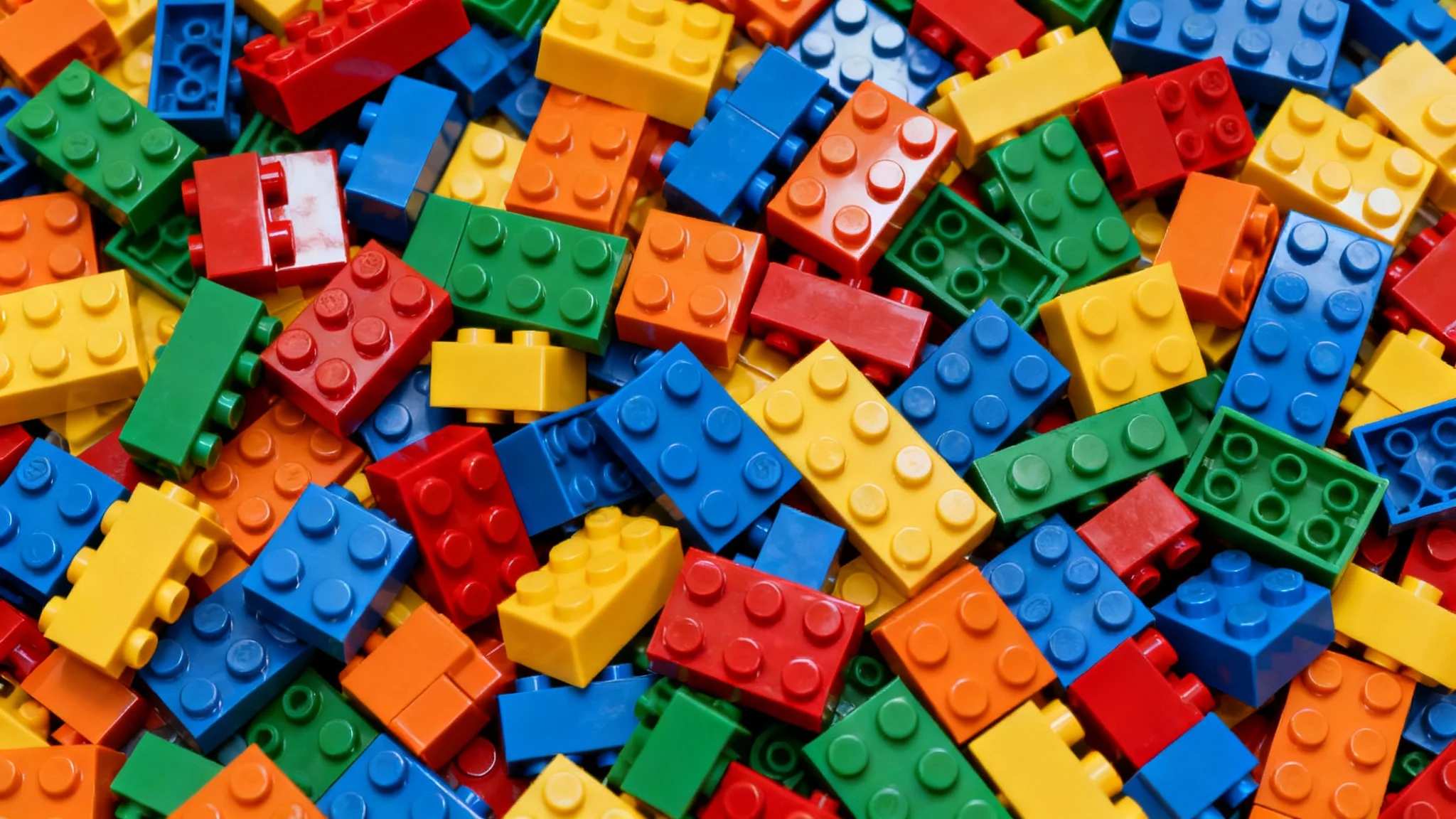 A top-down photograph showing a dense, colorful background made entirely of overlapping plastic toy bricks in various shapes and sizes.
