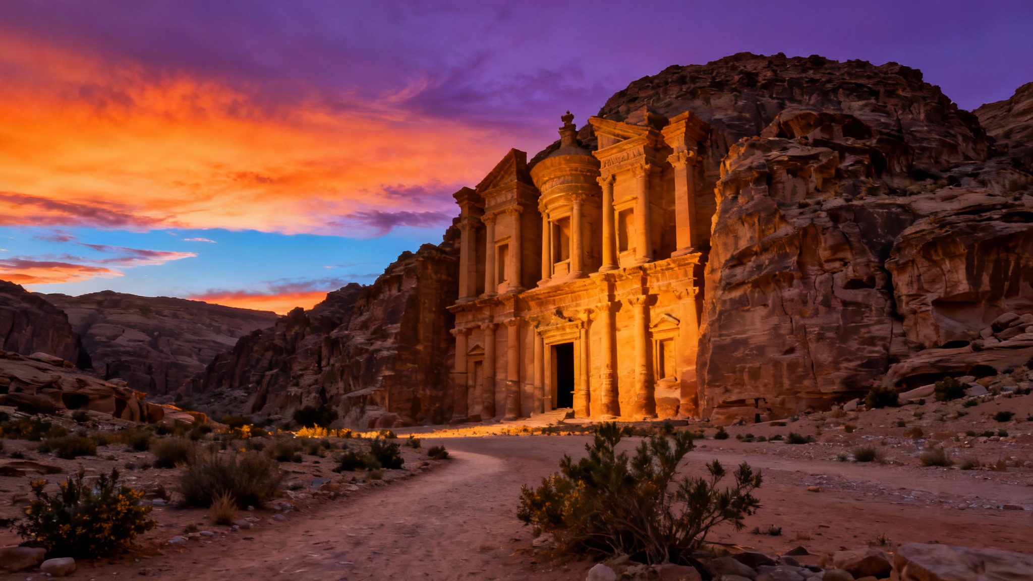 A stunning 16:9 wallpaper of the Petra Monastery in Jordan at sunset. The ancient rock-cut facade glows with warm, golden light from the setting sun. A dramatic sky with shades of orange and purple frames the scene, viewed from across a rugged desert landscape, creating a beautiful and epic final image.