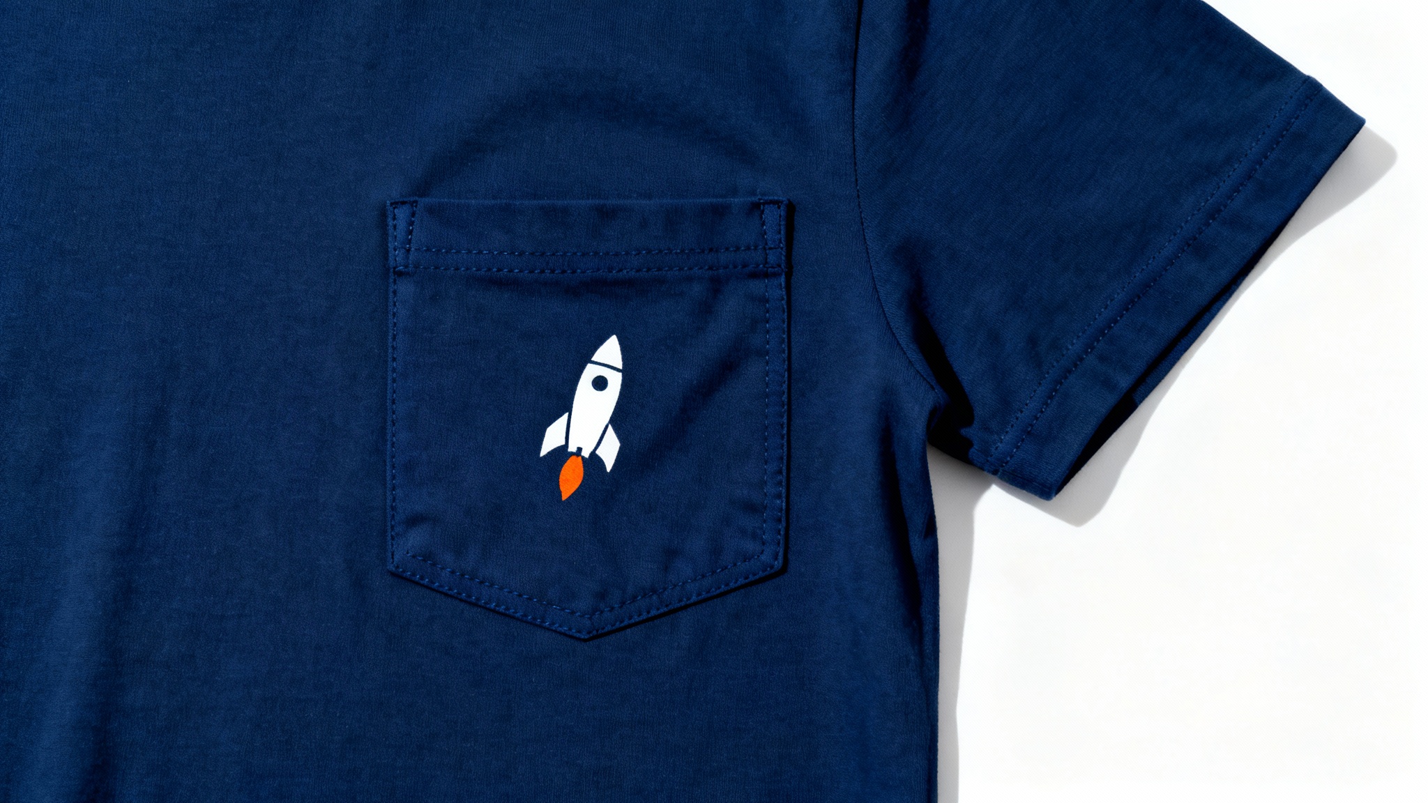 A close-up product shot of a small rocket ship graphic printed on the pocket of a navy blue t-shirt, set against a white background.