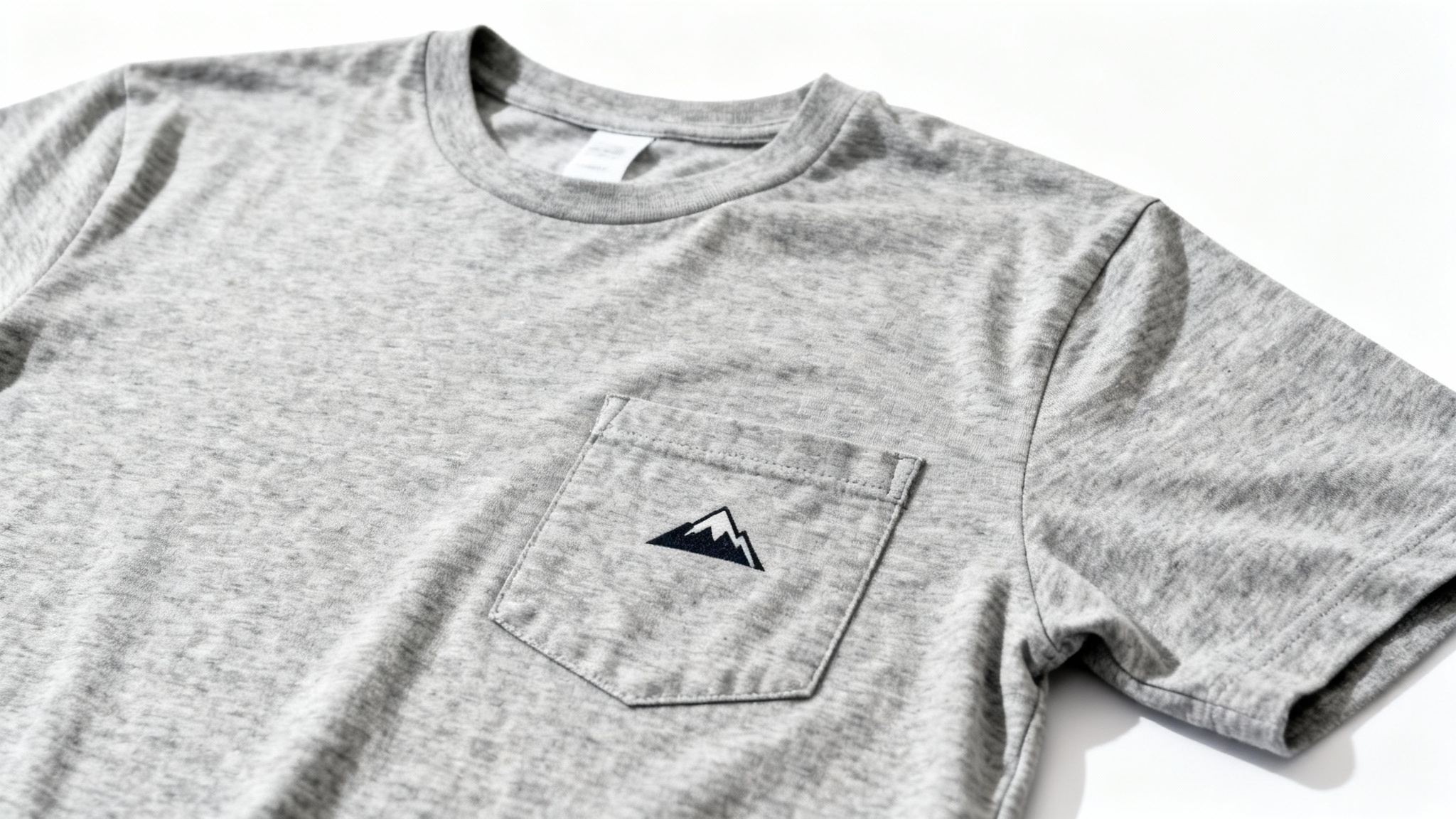 A close-up mockup of a small, minimalist mountain graphic printed perfectly on the pocket of a heather gray t-shirt, set against a clean white background.
