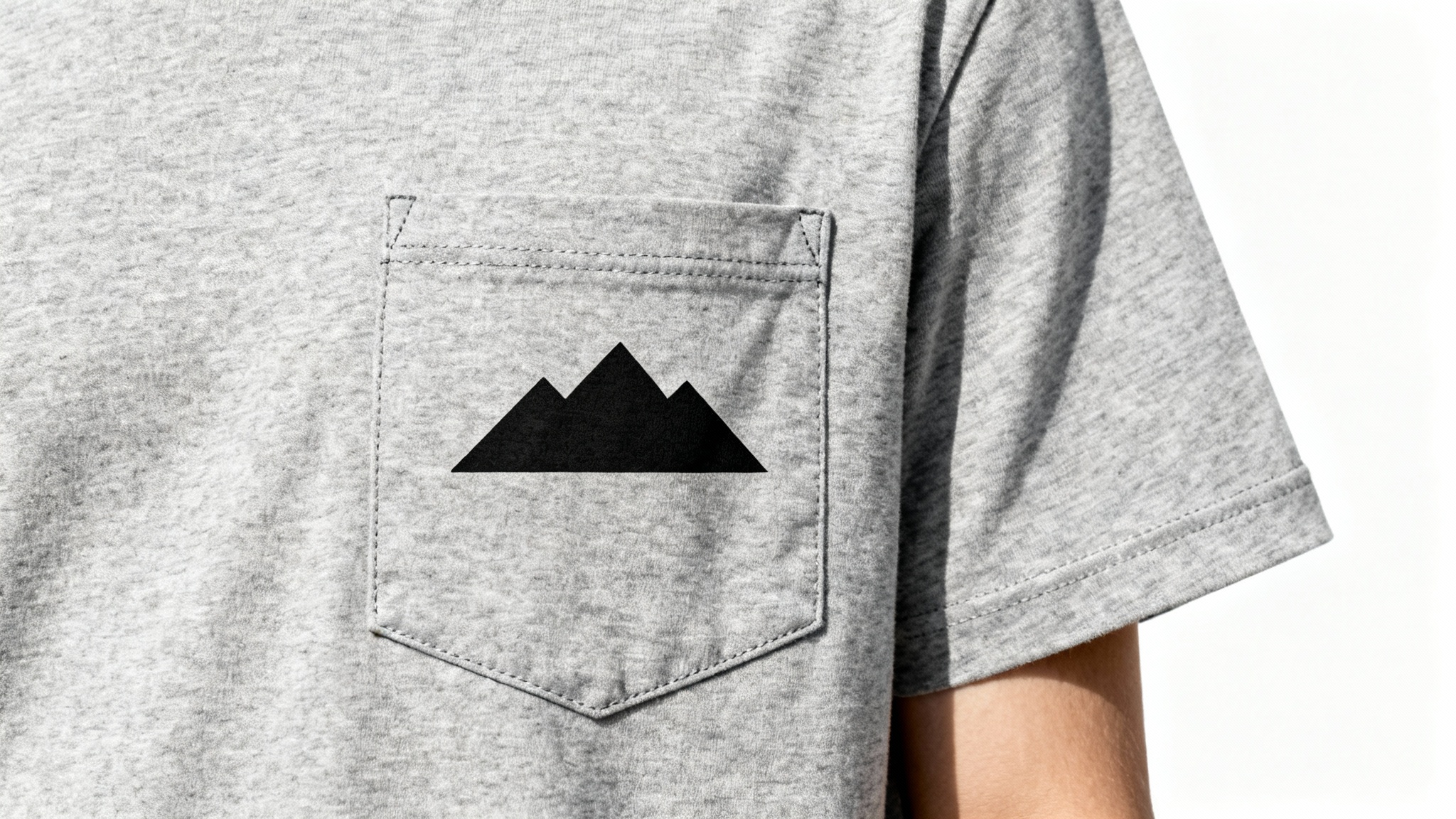 A detailed close-up shot of a heather gray t-shirt pocket with a small, minimalist black mountain range graphic printed on it, set against a clean white background.
