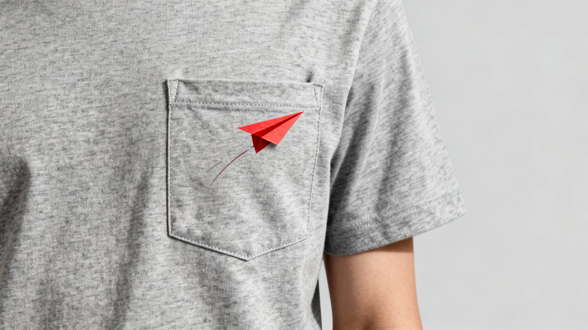 A close-up product photo of a heather grey t-shirt pocket featuring a small, minimalist red paper airplane graphic, on a clean light grey background.