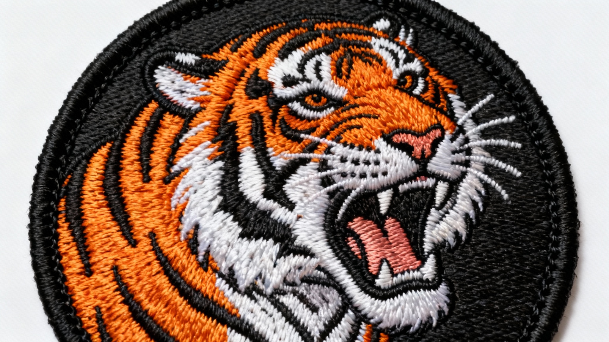A detailed, circular embroidered patch of a roaring tiger's head, showcasing intricate threadwork and vibrant colors, isolated on a plain white background.