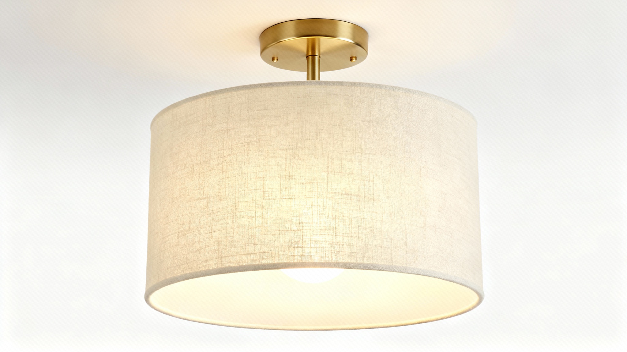 A modern semi-flush drum light with a cream linen shade and satin brass base, shown illuminated against a clean white background.