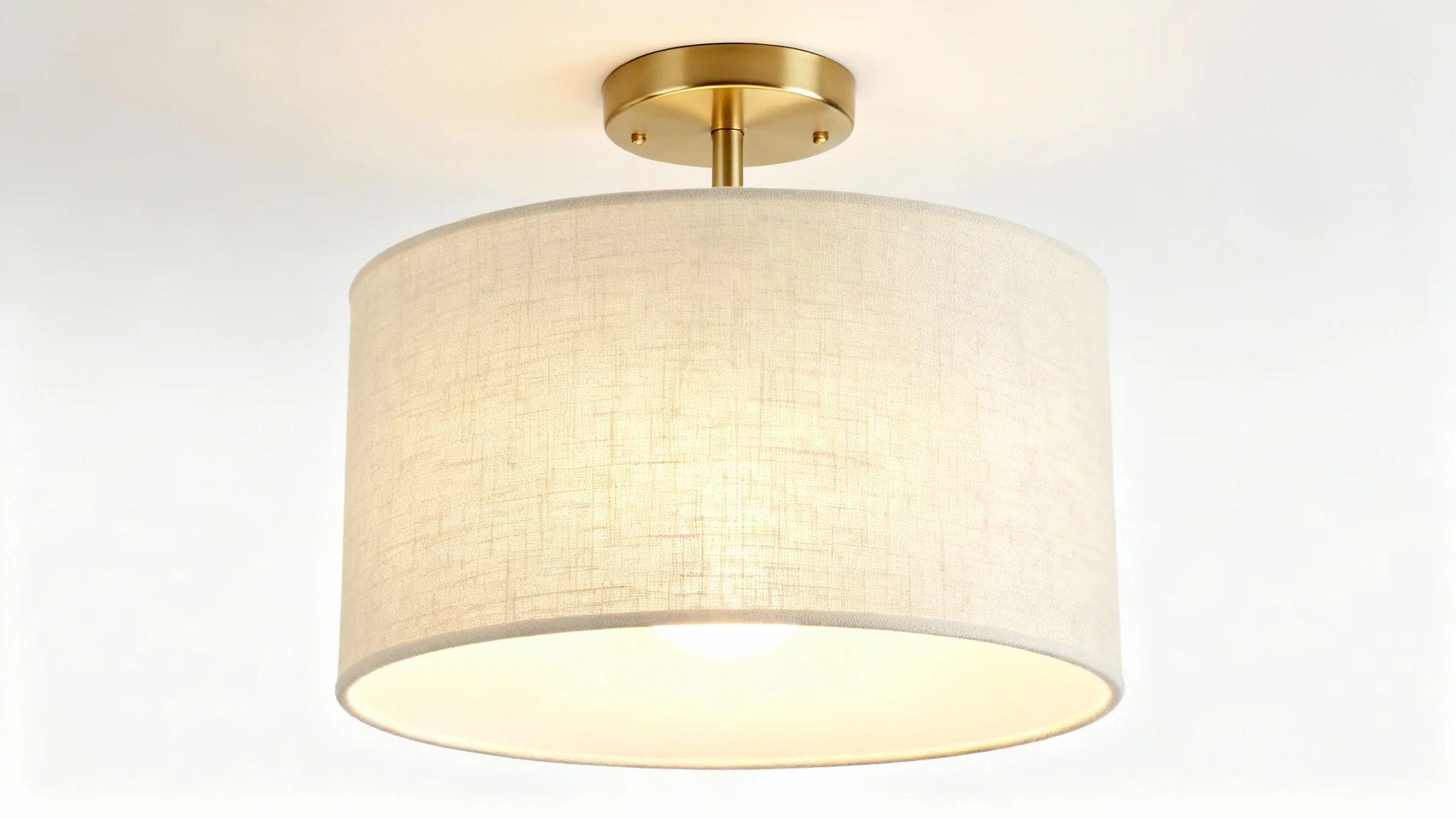 A modern semi-flush drum light with a cream linen shade and satin brass base, shown illuminated against a clean white background.