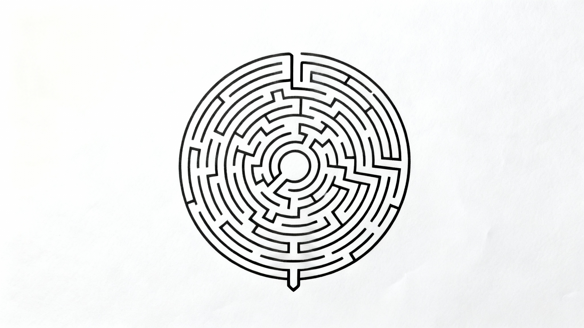 A black and white circular maze on a plain white background, featuring a complex pattern of paths from an entrance to an exit.